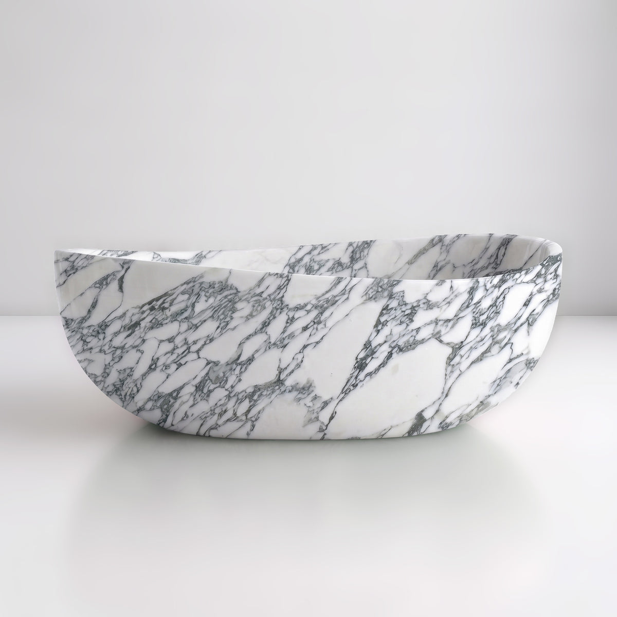 Front studio view of the freestanding Arabescato marble wave bathtub, showcasing the organic dipping rim silhouette and dramatic charcoal-grey breccia veining across the white stone exterior.
