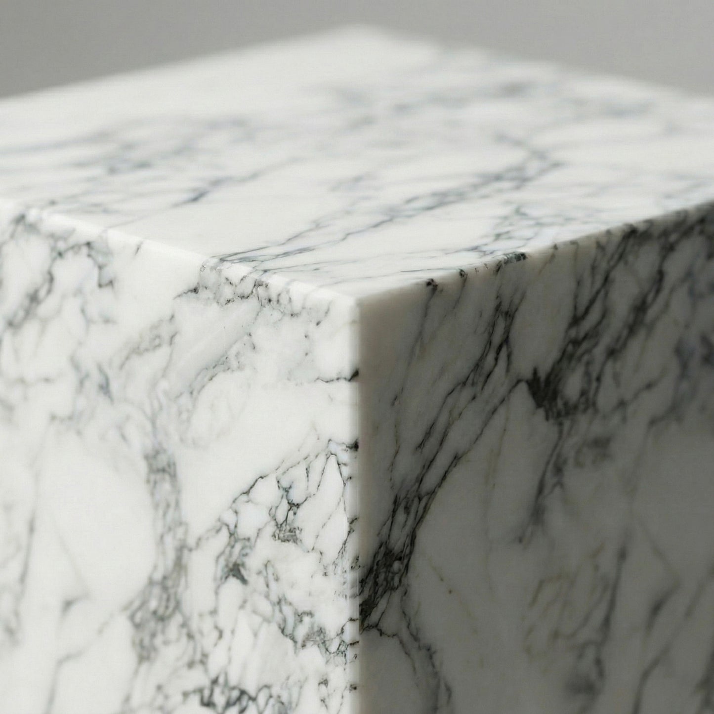 An extreme close-up of the corner of an Arabescato marble pedestal, highlighting the distinctive dark gray vein loops and the sharp, seamless mitered edge of the polished stone.
