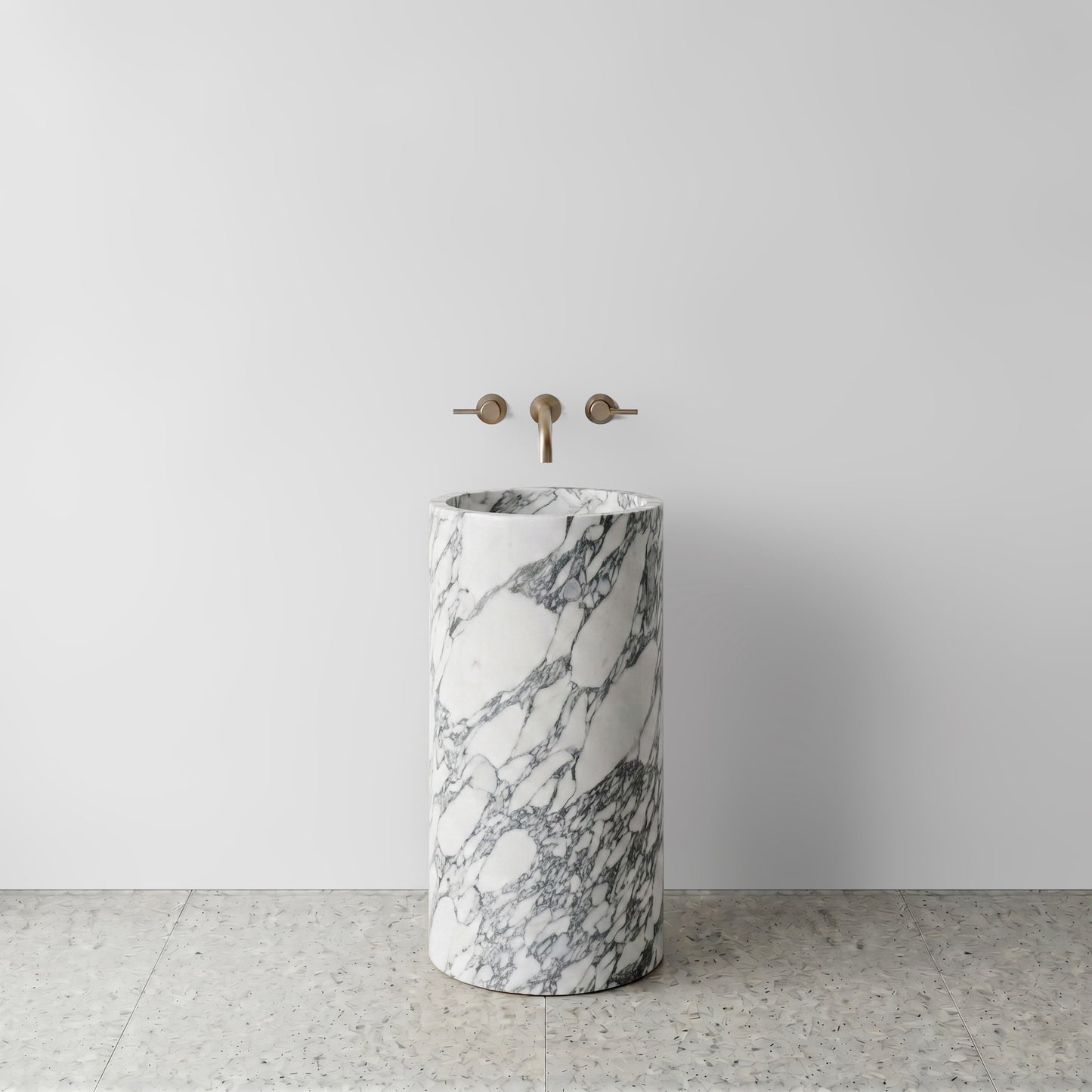 Front view of a monolithic Arabescato marble cylinder pedestal sink on light terrazzo flooring with wall-mounted brushed bronze tapware.