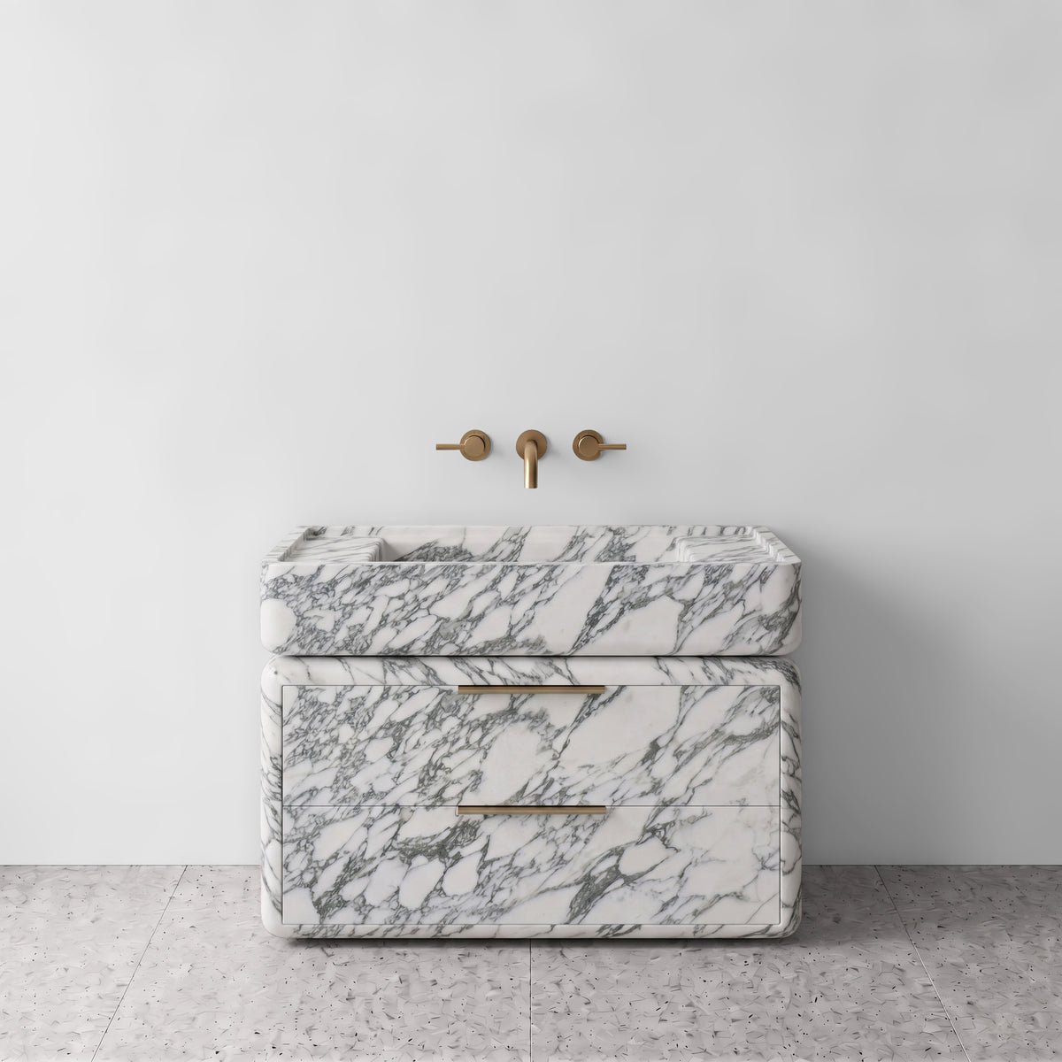 Front studio view of the freestanding Arabescato marble double-drawer vanity, featuring rounded corners, an integrated trough basin, brushed brass linear pulls, and wall-mounted three-hole brass tapware set against a white wall and terrazzo flooring.