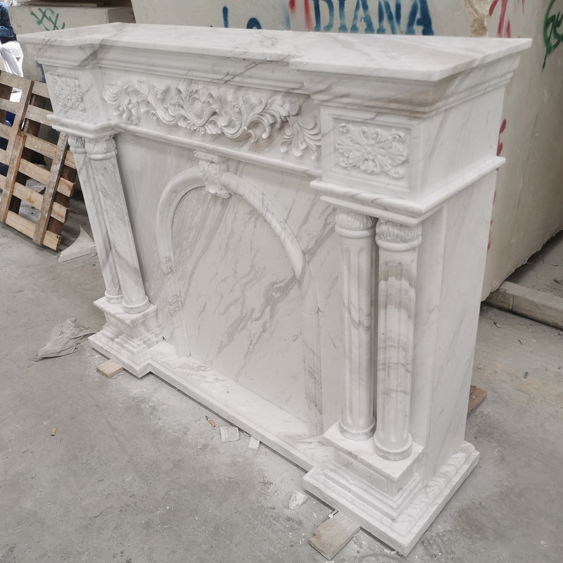 A classically designed, hand-carved white marble fireplace surround featuring ornate columns and floral relief details, demonstrating GustavDesign’s expertise in bespoke stone masonry.