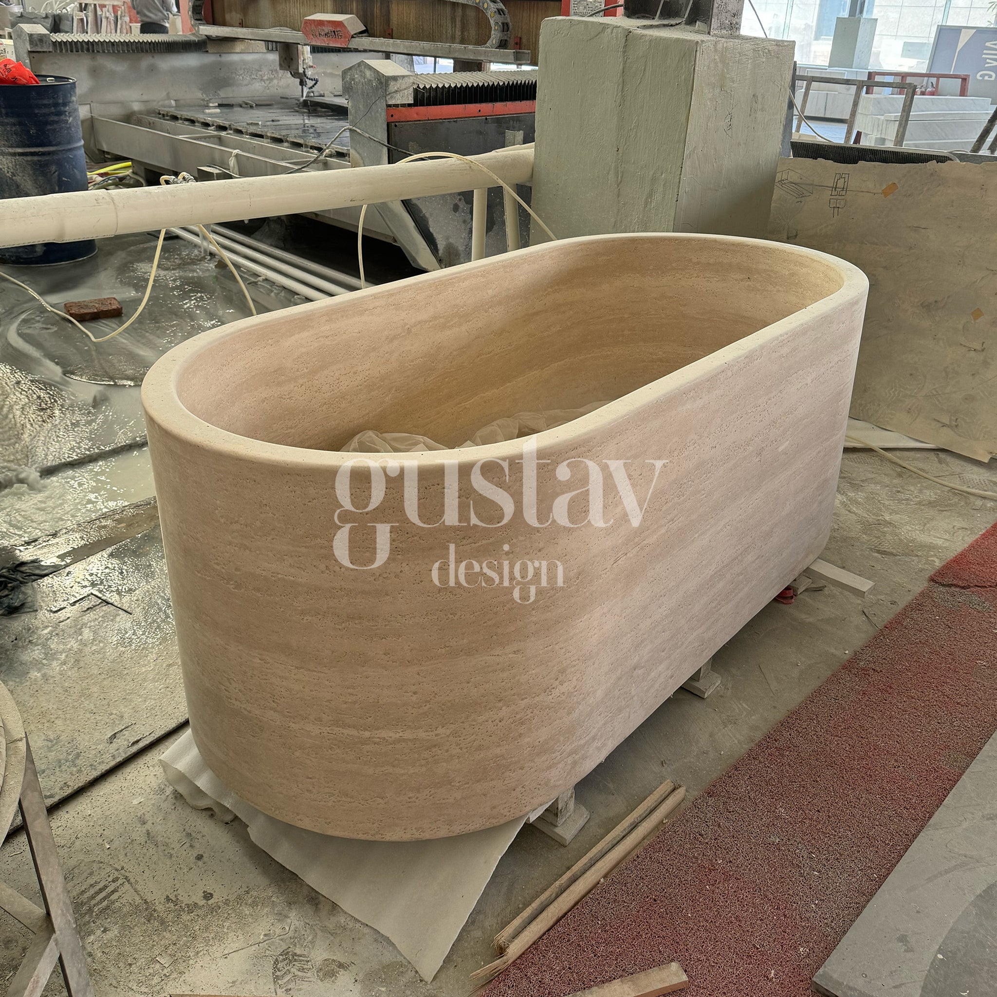 Hand-carved oval beige travertine bathtub in a fabrication workshop, luxury natural stone soaking tub by Gustav Design.