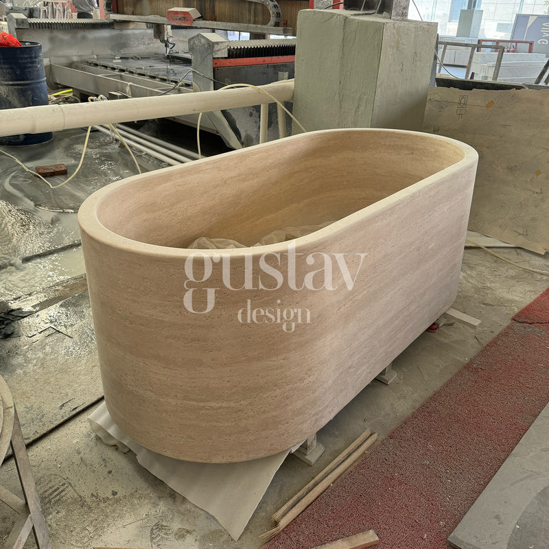 Hand-carved oval beige travertine bathtub in a fabrication workshop, luxury natural stone soaking tub by Gustav Design.