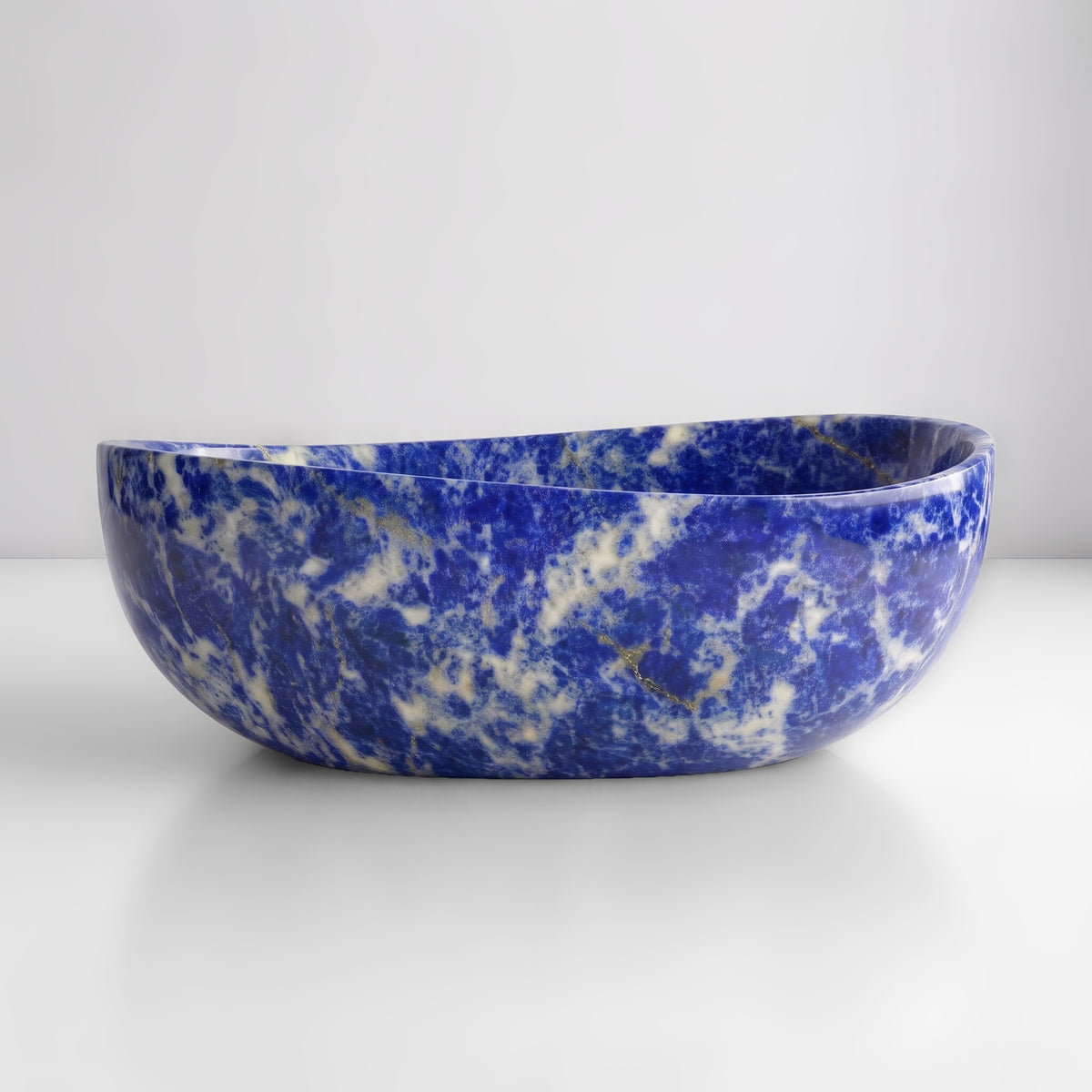 Front studio view of the freestanding Blue Sodalite marble wave bathtub, showcasing the organic dipping rim silhouette and vivid cobalt-blue ground with white calcite clouding across the stone exterior.