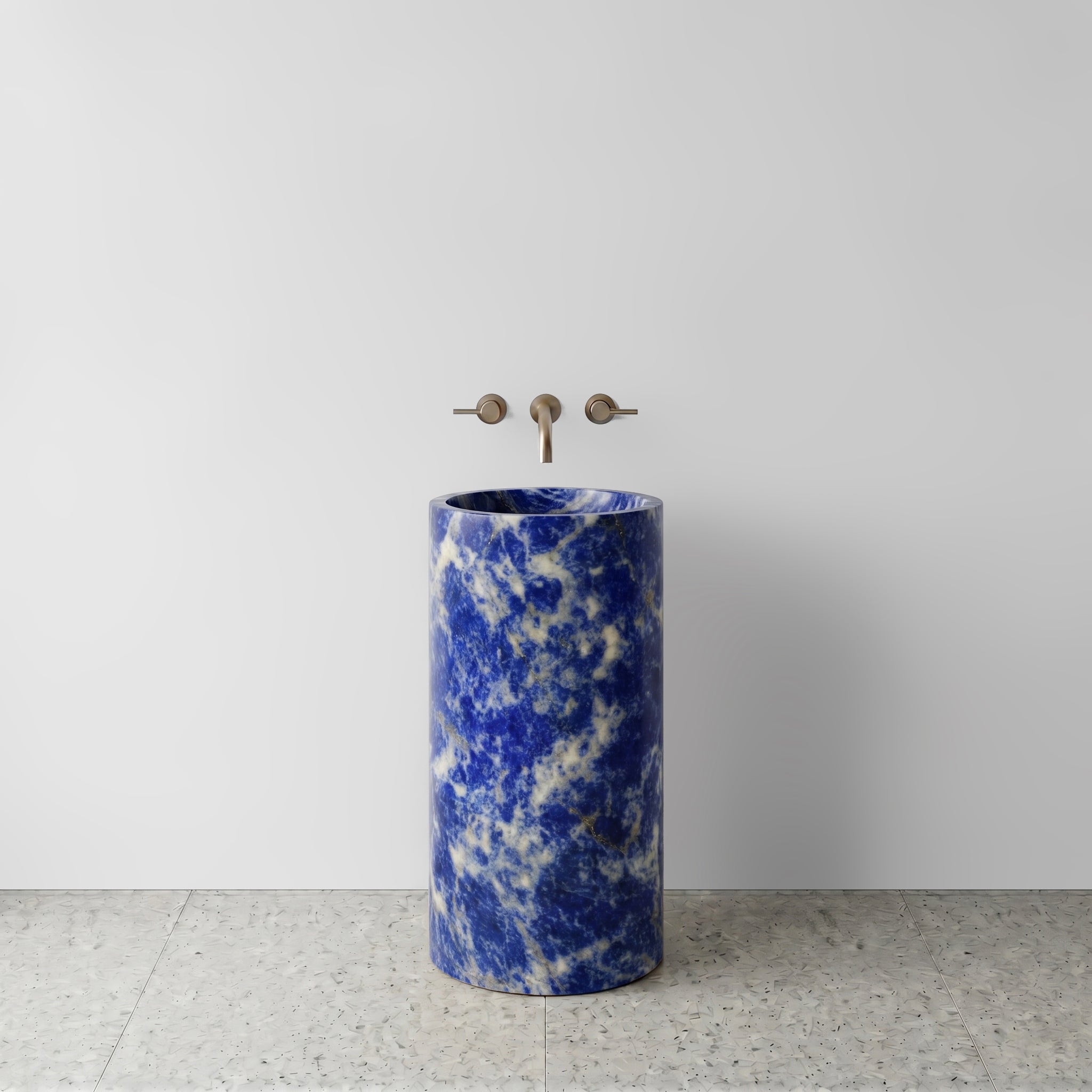 Front view of a monolithic Blue Sodalite cylinder pedestal sink on light terrazzo flooring with wall-mounted brushed bronze tapware.