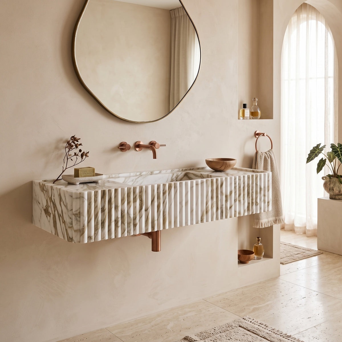 Bright, warm-minimalist bathroom interior featuring the floating Calacatta Gold fluted stone sink paired with a round brass mirror, textured plaster walls, and an arched window.