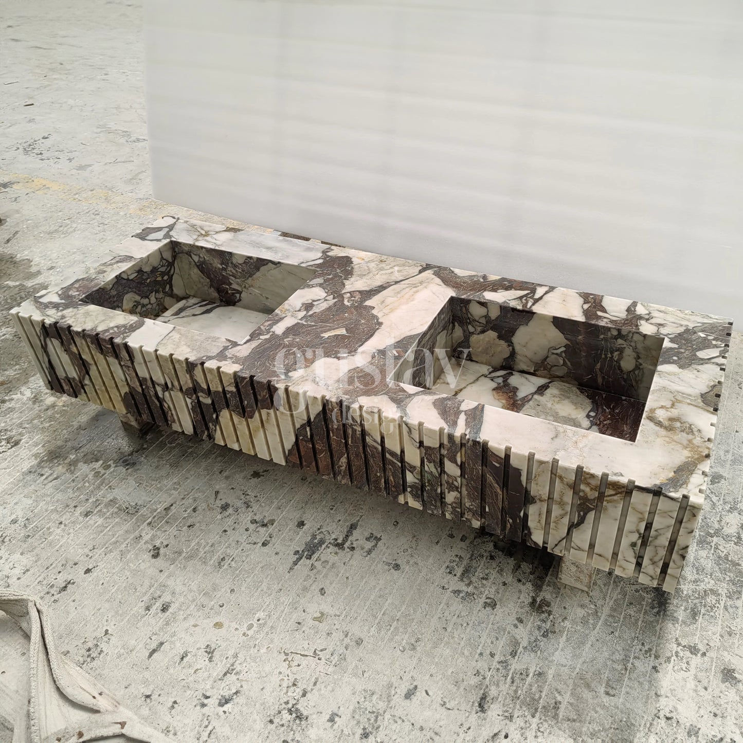 A high-angle, front-facing view of a double-basin floating vanity carved from Calacatta Viola marble. The stone features heavy, dramatic veining in shades of deep burgundy and plum against a creamy white background. The front and sides of the vanity are designed with a fluted, vertical ribbed texture, creating a rhythmic architectural look. The unit is shown in a workshop setting on a concrete floor.