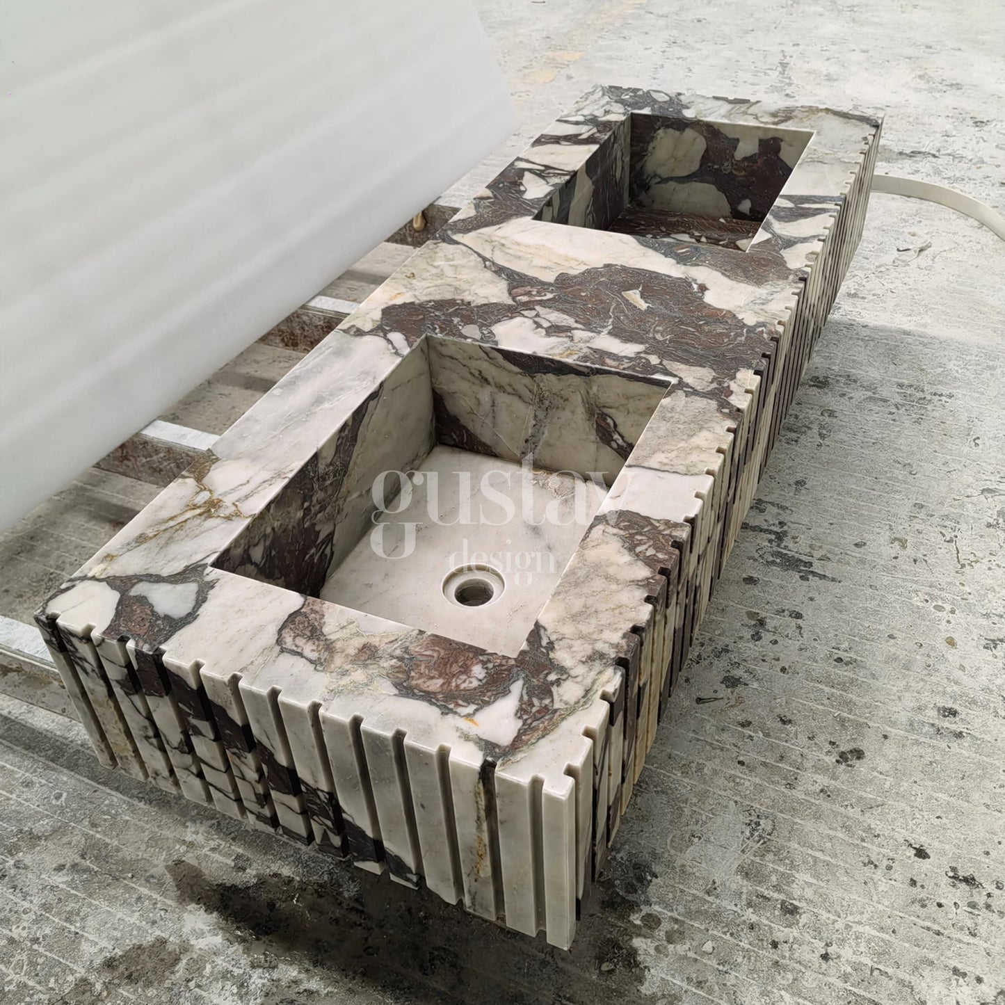 A side-angle view of the marble vanity, highlighting the depth of the rectangular basins and the precision of the fluted edges. The integrated marble sinks show continuous veining from the countertop into the basin walls. A drain hole is visible at the bottom of the right-hand sink. The vanity is supported by temporary metal frames in a fabrication warehouse.
