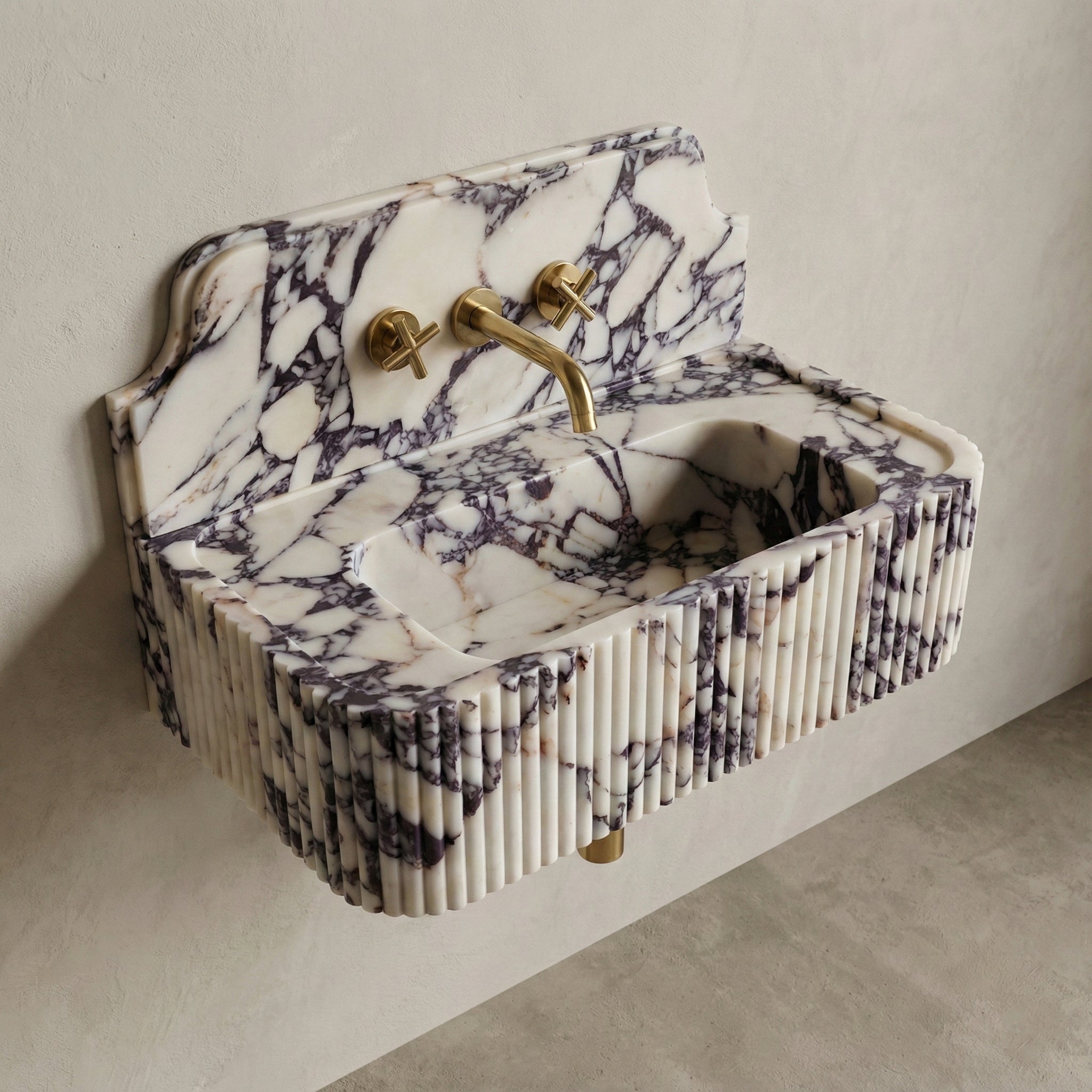 High-angle side view of a vintage-inspired Calacatta Viola marble sink, featuring a curved stone backsplash, fluted apron, and brass cross-handle faucet.