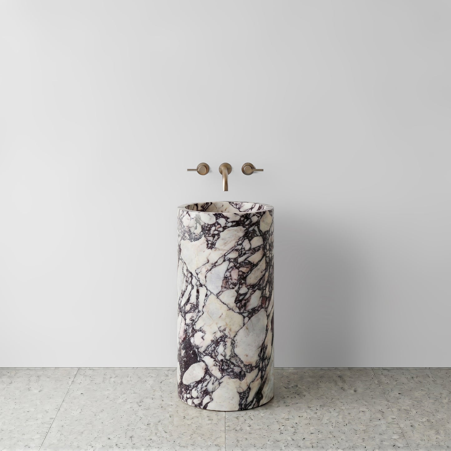 Front view of a monolithic Calacatta Viola marble cylinder pedestal sink on light terrazzo flooring with wall-mounted brushed bronze faucet.