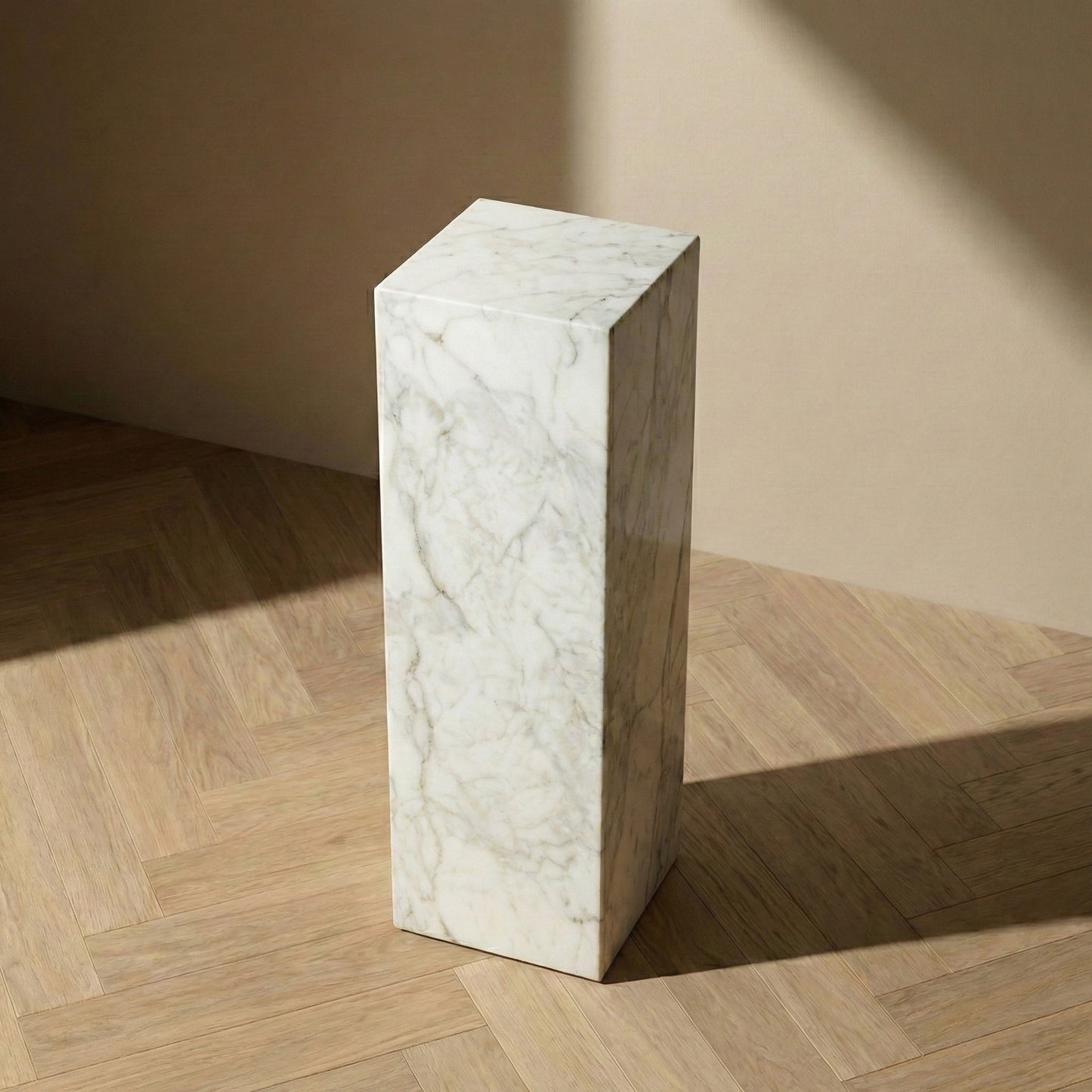 An elevated perspective view of a tall, rectangular Carrara marble plinth on light oak herringbone flooring, highlighting its clean architectural lines and natural stone patterns.