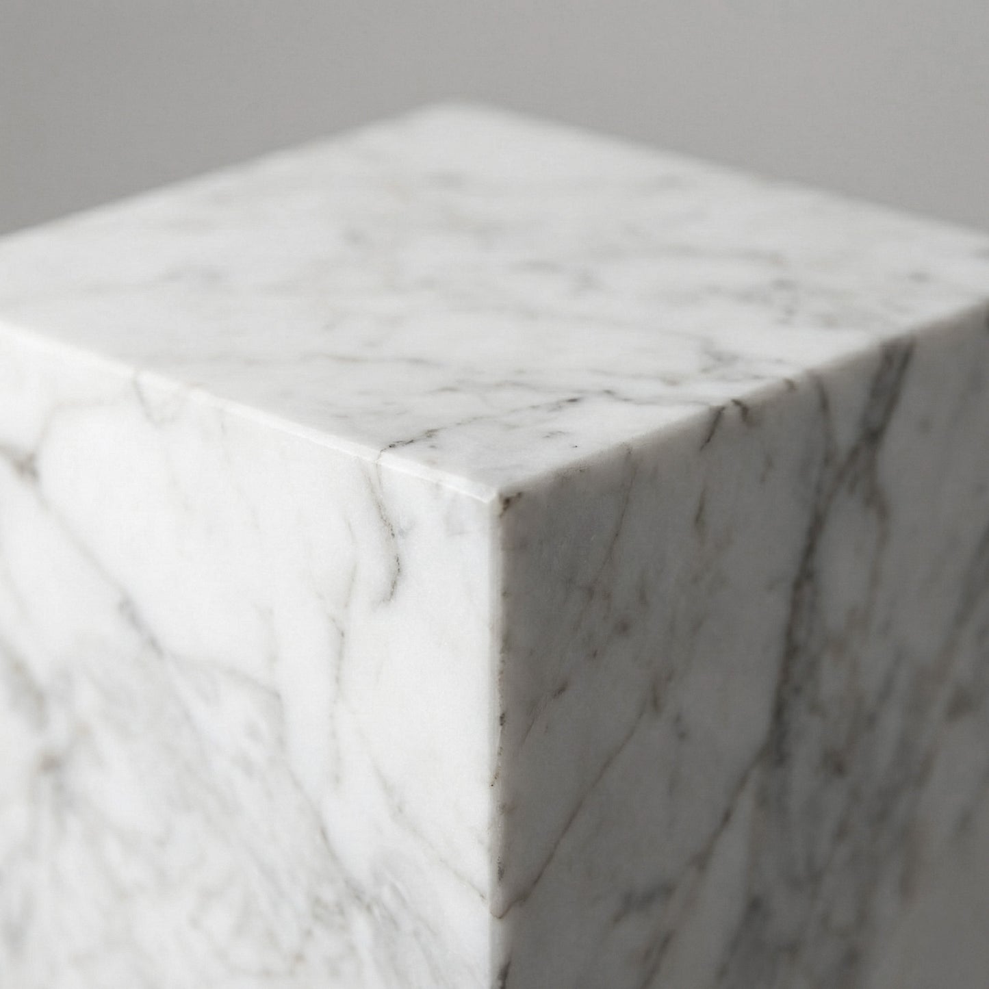 A macro shot of the corner of a White Carrara marble pedestal, showing the crisp mitered edge and the soft, smoky gray veins characteristic of authentic Italian marble.