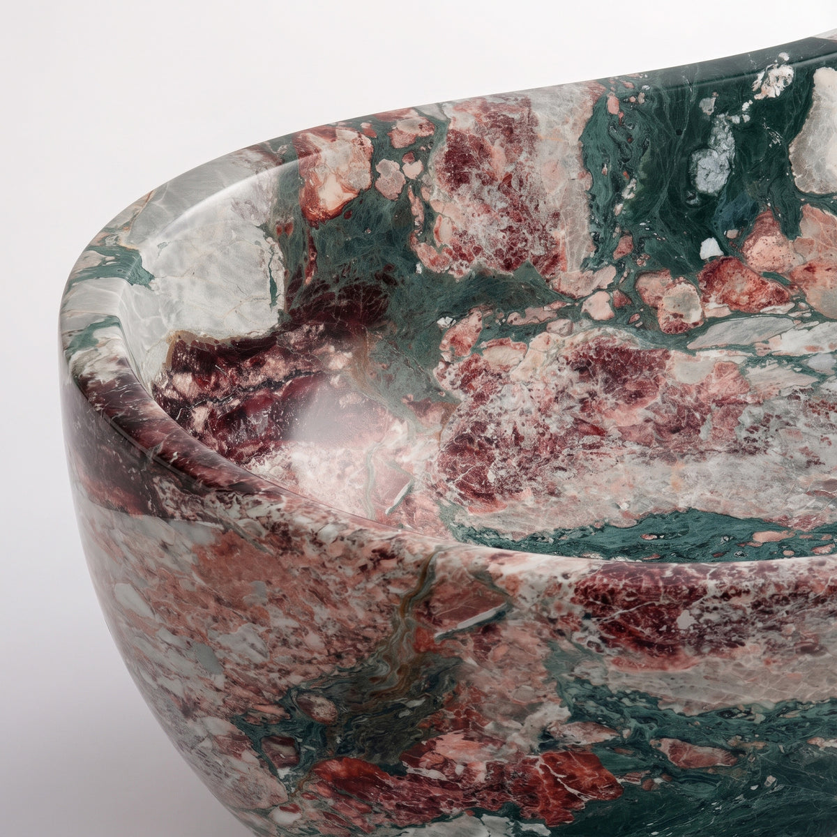 Close-up overhead detail shot of the Four Seasons marble wave bathtub rim, displaying the hand-finished curve, smooth stone surface, and high-contrast blend of deep claret red, sage, and dark green crystalline breccia.