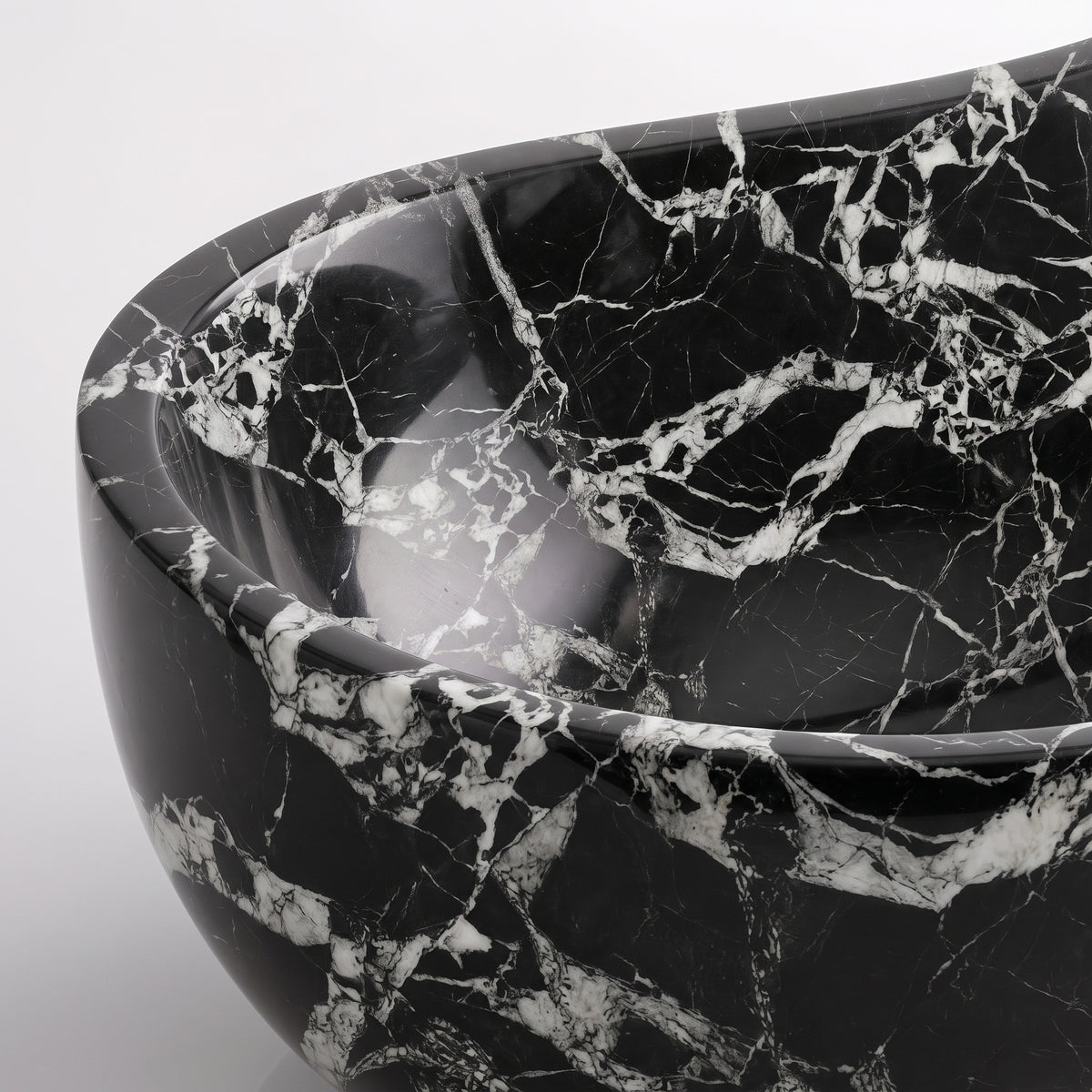 Close-up overhead detail shot of the Grand Antique marble wave bathtub rim, displaying the hand-finished curve, polished stone texture, and sharp contrast between fractured white calcite clasts and deep obsidian marble.