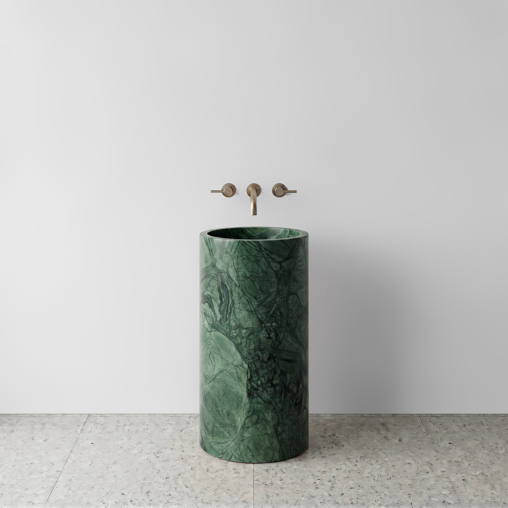 Front view of a solid Green Guatemala marble cylinder pedestal sink on light terrazzo flooring with wall-mounted brushed bronze faucet.