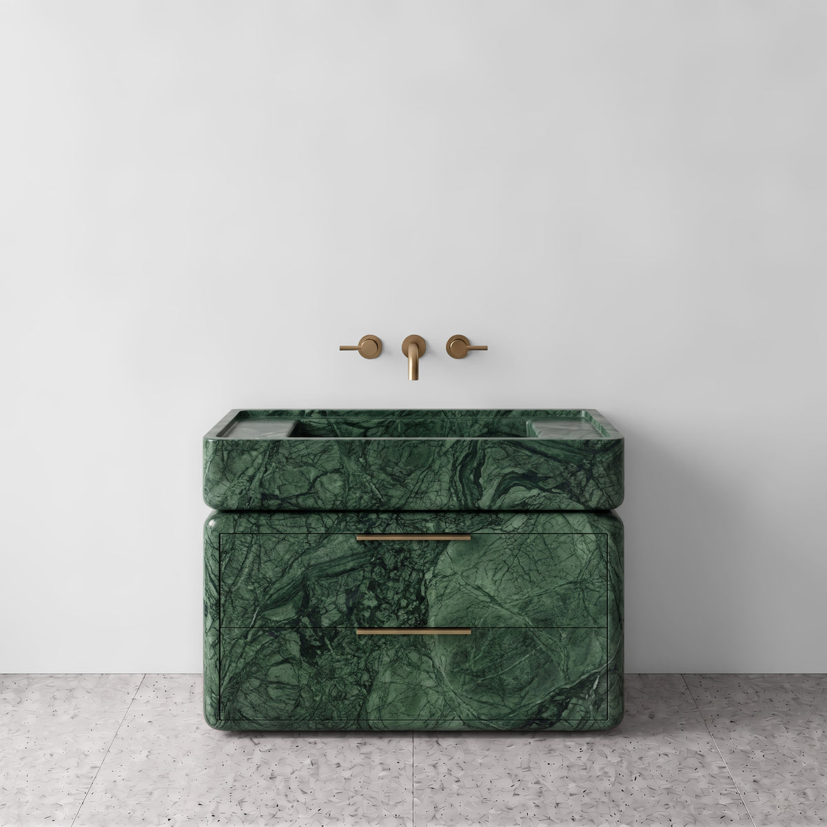 Front studio view of the freestanding Green Guatemala marble double-drawer vanity, featuring rounded corners, an integrated trough basin, horizontal brushed brass pulls, and wall-mounted three-hole brass tapware set against a white wall and terrazzo flooring.