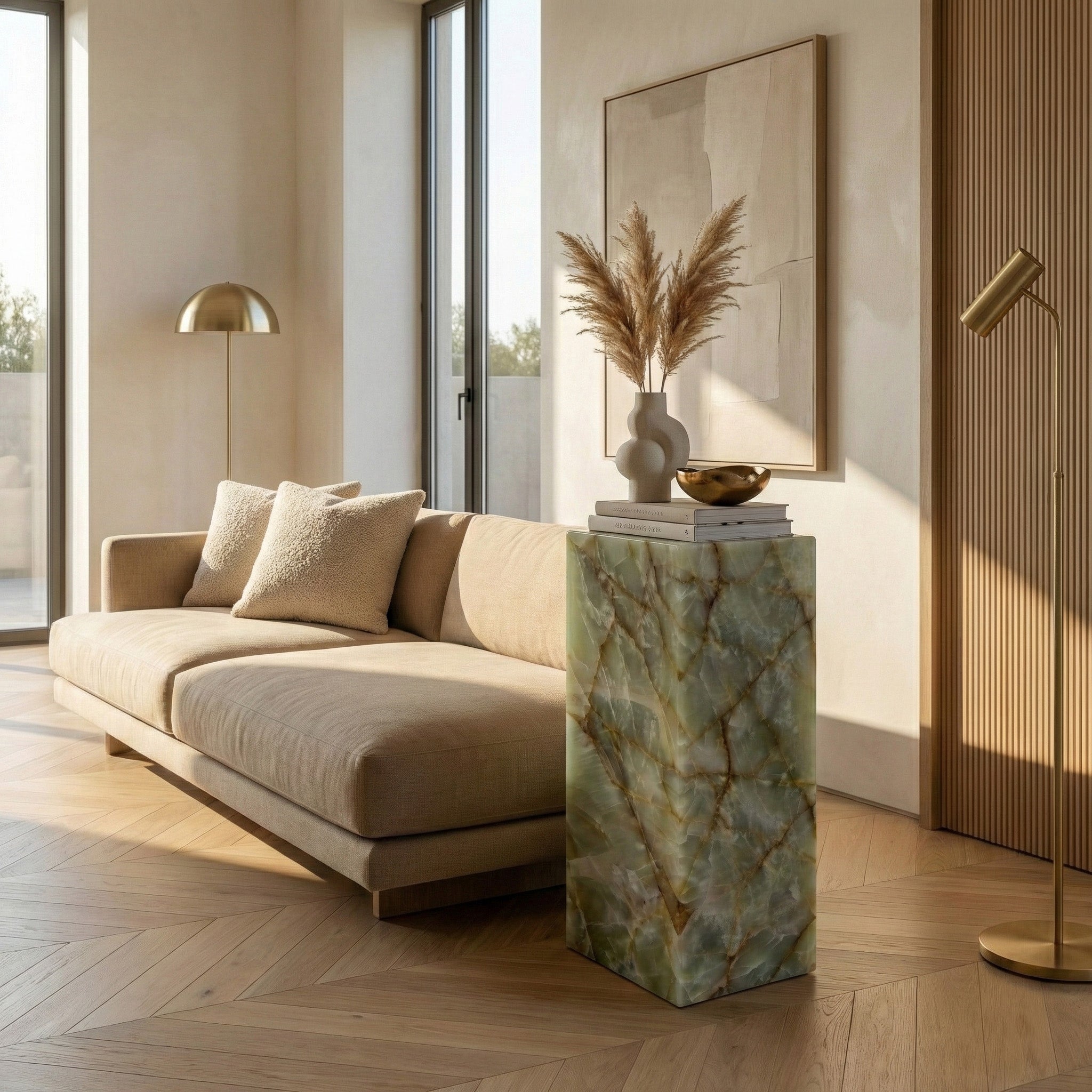 A modern, sun-drenched living room featuring a Green Onyx pedestal stand used as a display for a vase with pampas grass, positioned next to a tan sofa and minimalist gold lighting.