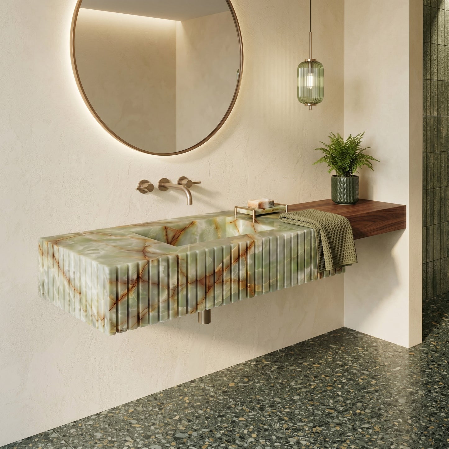 Warm minimalist bathroom interior featuring a fluted green onyx single sink integrated with a floating walnut wood shelf, a round brass mirror, a fluted green glass pendant light, and dark terrestrial terrazzo floor tiles.