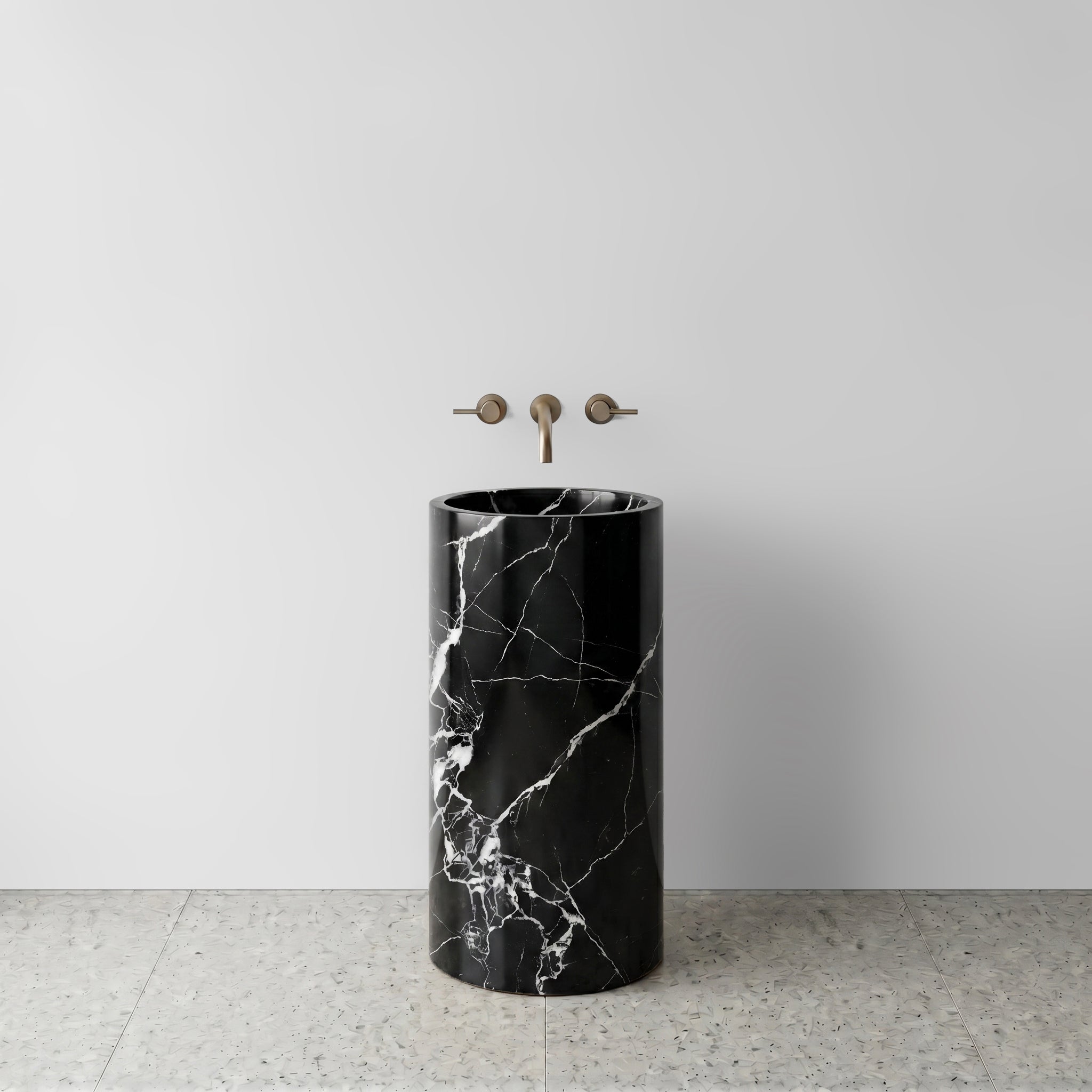 Front view of a solid Nero Marquina black marble cylinder pedestal sink on light terrazzo flooring with wall-mounted bronze tapware.