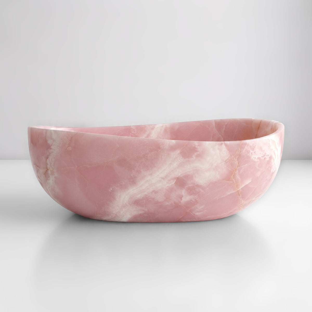 Front studio view of the freestanding Pink Onyx wave bathtub, showcasing the organic dipping rim silhouette and soft blush-pink crystalline field with translucent white clouding across the polished stone exterior.