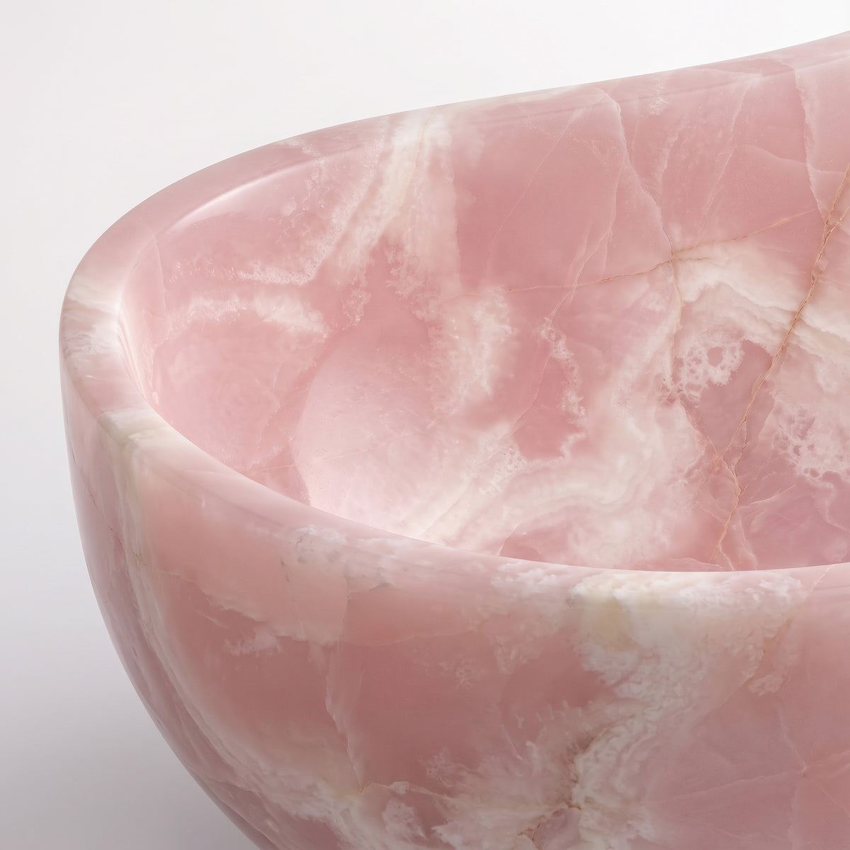 Close-up overhead detail shot of the Pink Onyx wave bathtub rim, displaying the hand-finished curve, polished stone translucency, and soft pastel pink clouding accented by fine white crystalline veining.