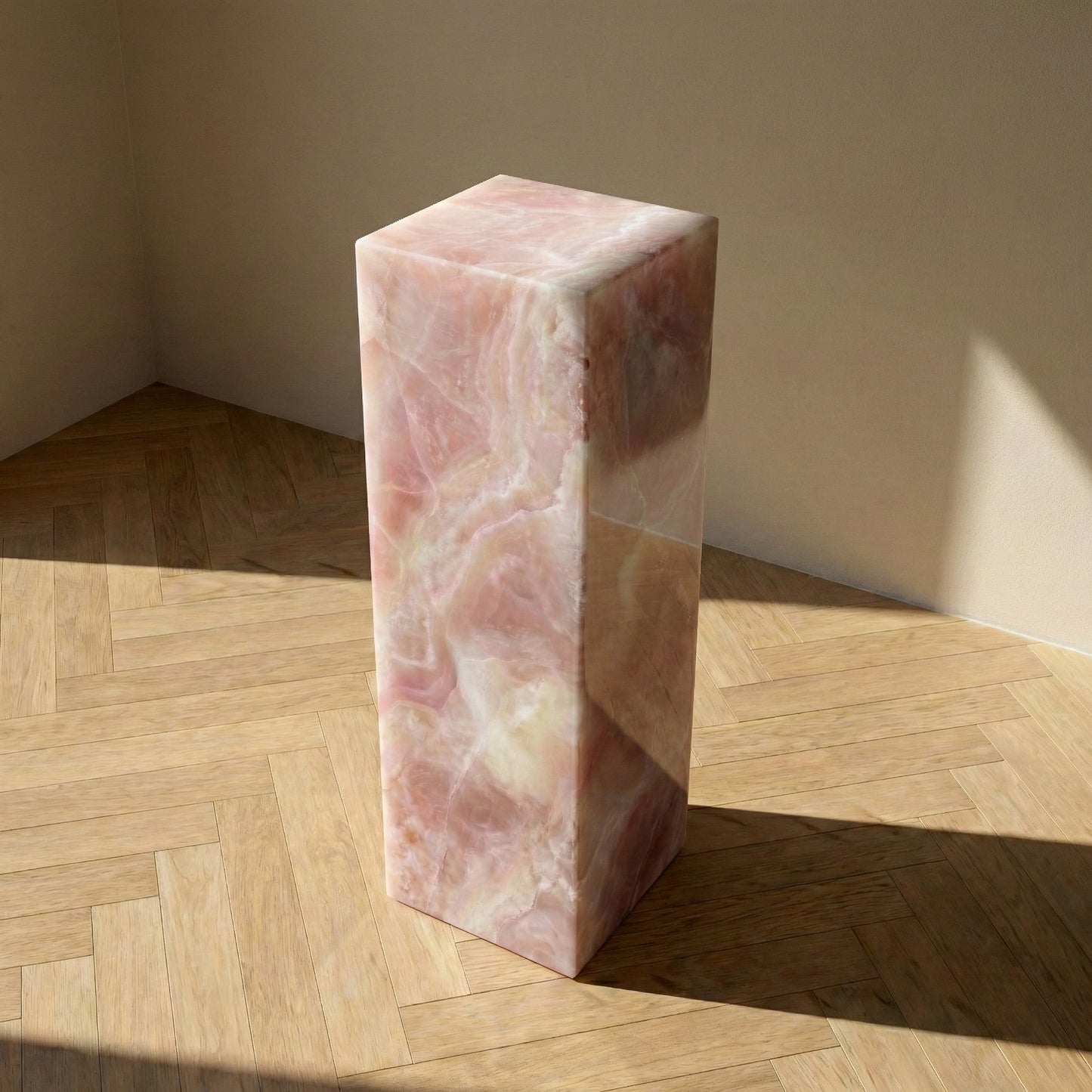 An elevated perspective of a rectangular Pink Onyx plinth on a wood herringbone floor, highlighting the stone's natural swirling patterns and high-gloss polished surface.