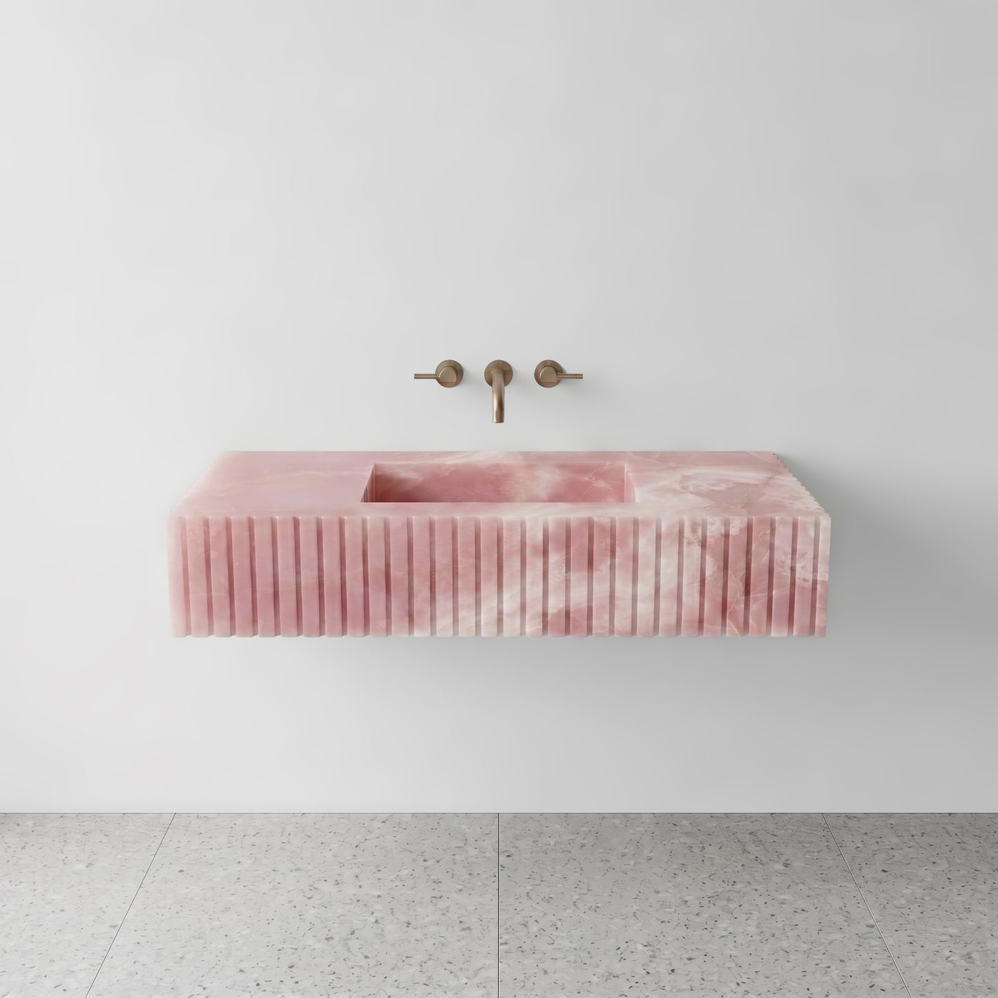 Front studio view of a luxury floating single sink vanity made of translucent Pink Onyx, featuring an architectural fluted apron front design and a wall-mounted brushed bronze faucet.