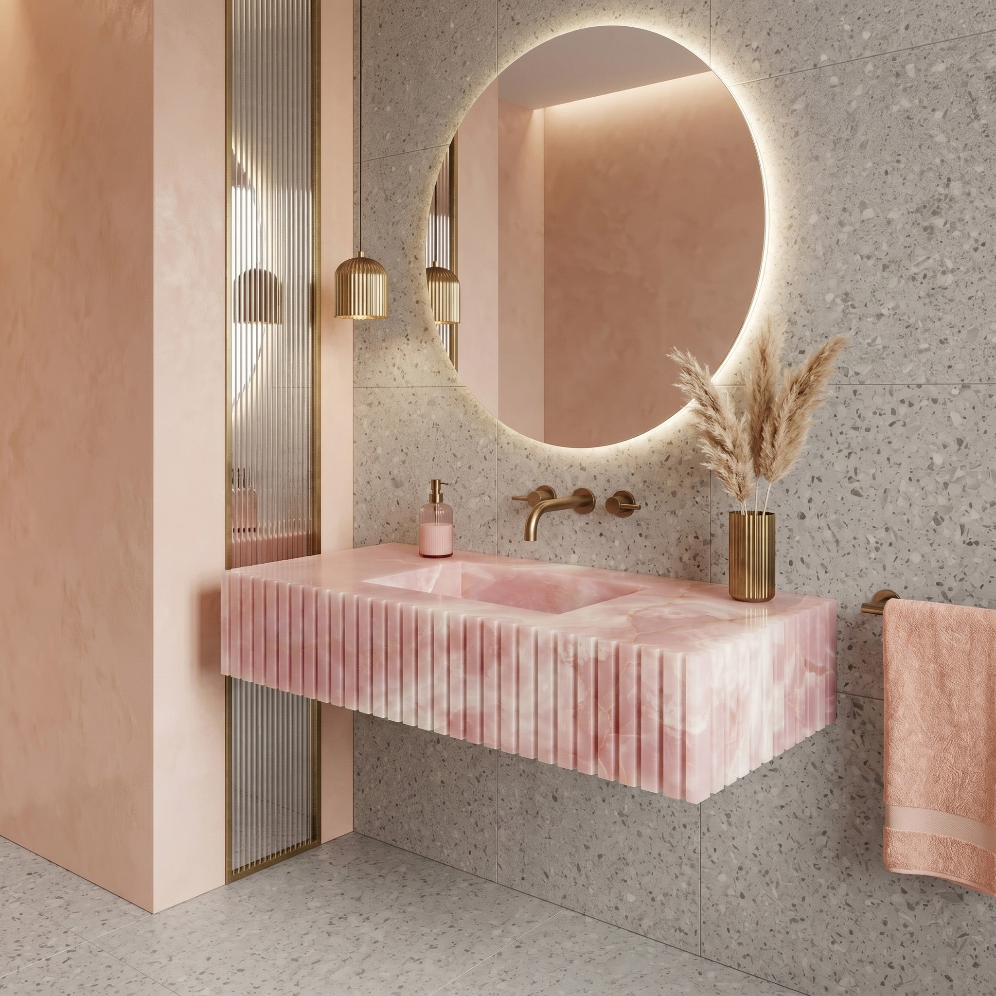 Modern pastel bathroom interior featuring a fluted pink onyx single washbasin, grey terrazzo wall and floor tiles, a large round backlit mirror, a reeded glass insert, and warm gold accents.