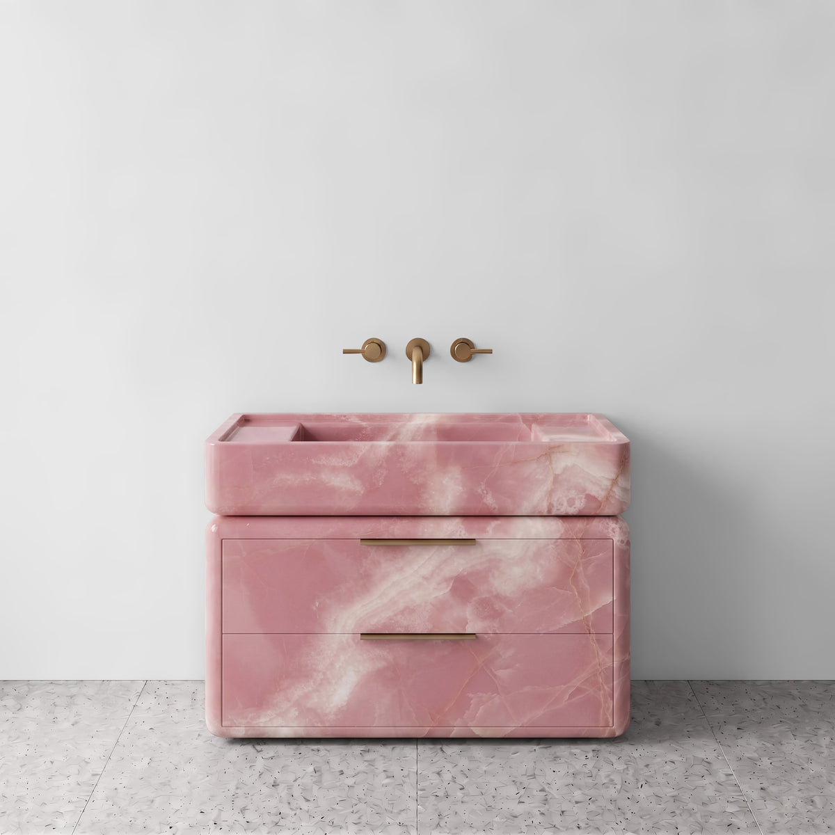 Front studio view of the freestanding Pink Onyx double-drawer vanity, featuring rounded corners, an integrated trough basin, horizontal brushed brass pulls, and wall-mounted three-hole brass tapware set against a white wall and terrazzo flooring.
