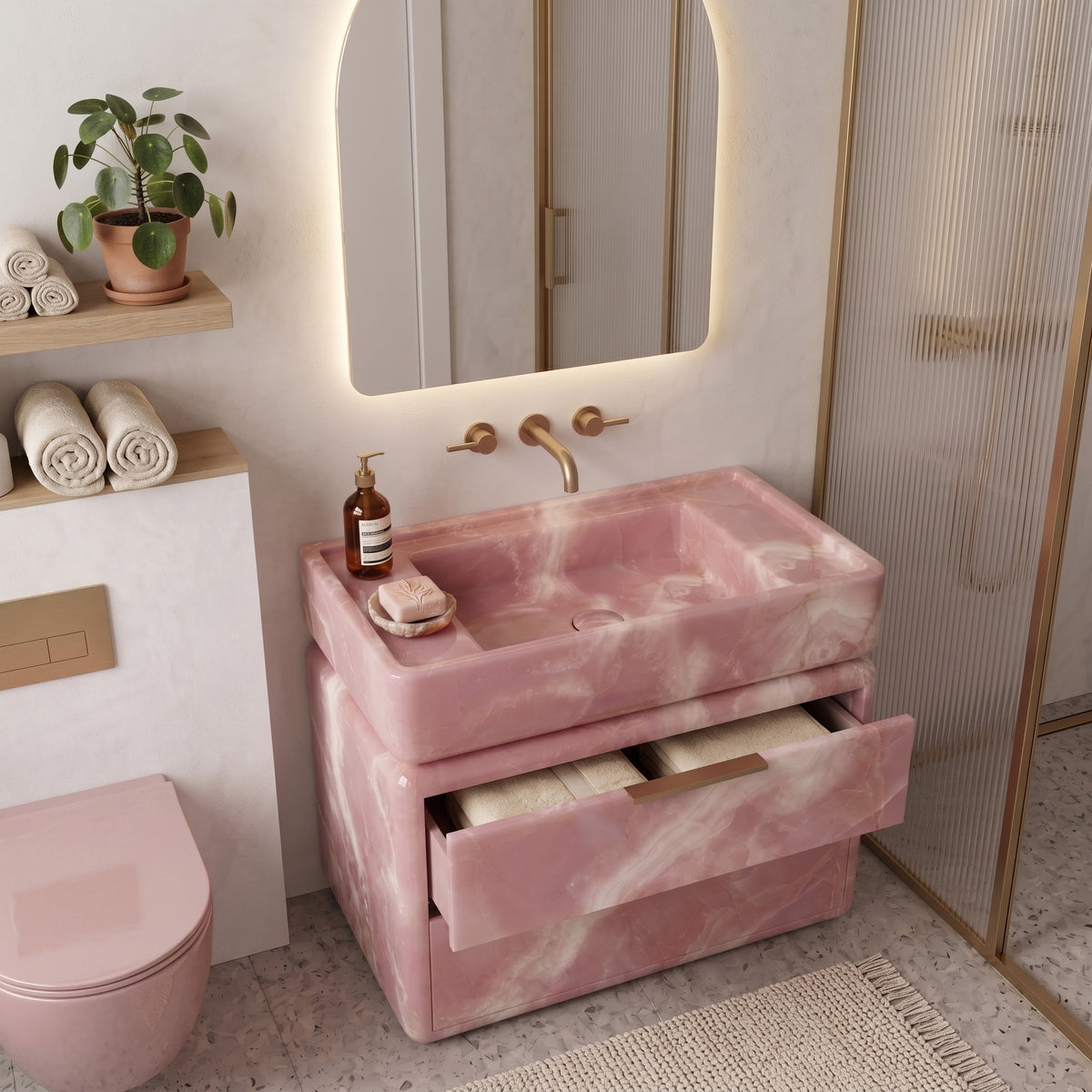 Luxury pastel bathroom interior featuring the curved Pink Onyx double-drawer vanity with an open top drawer displaying folded towels, paired with wall-mounted brushed brass tapware, an arched backlit mirror, a matching pink wall-hung toilet, floating timber shelving, and a fluted glass shower screen on terrazzo tile.