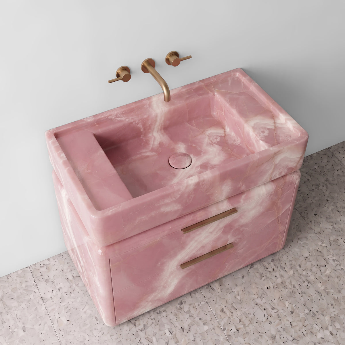 High-angle perspective view of the Pink Onyx double-drawer vanity, highlighting the integrated trough sink with a matching stone drain cover, raised perimeter border, rounded drawer corners, brushed brass linear hardware, and luminous blush-pink crystalline mineral movement.
