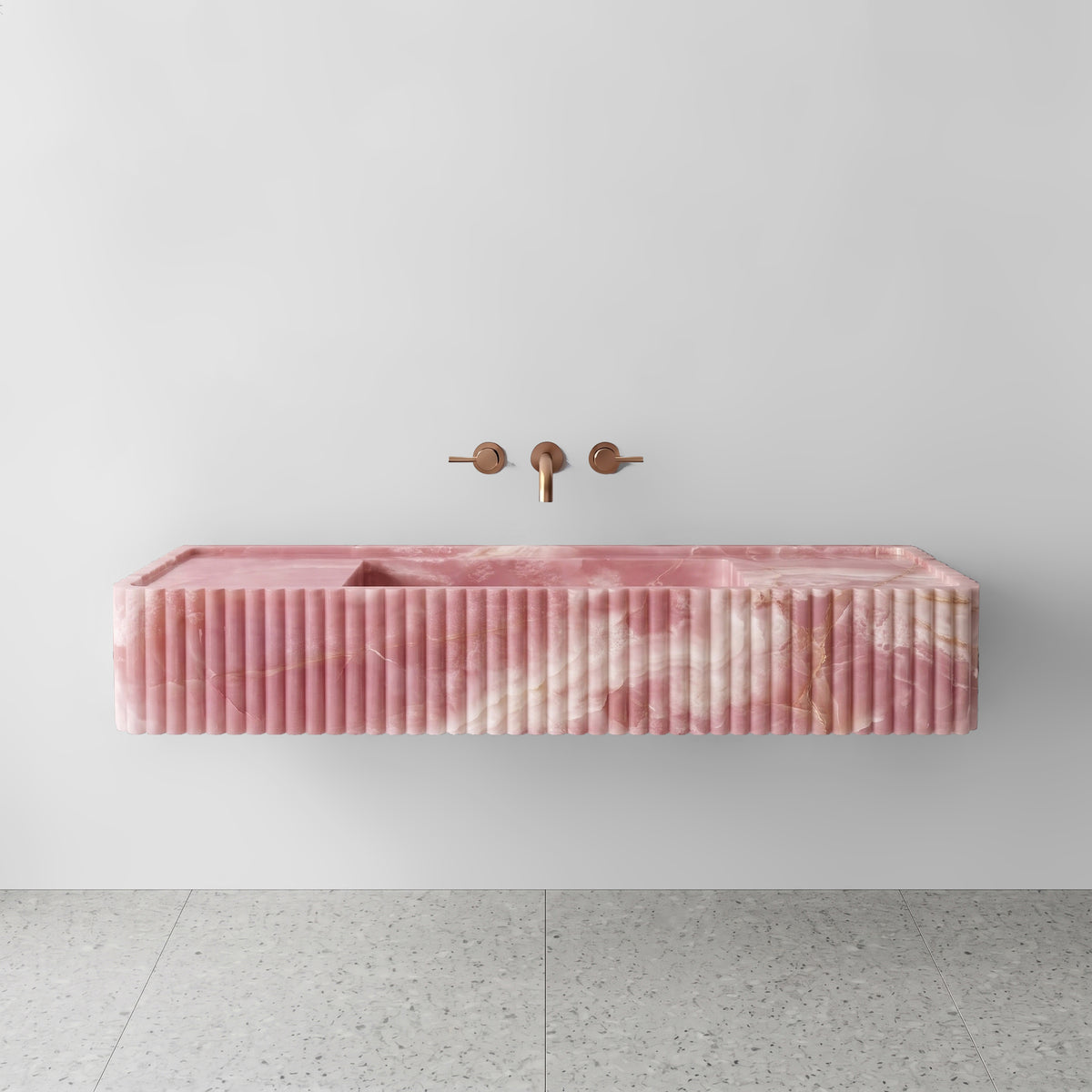 Front view of the Pink Onyx fluted monolith sink showing translucent blush and crystalline ivory banding across the vertical convex ribbed apron and wall-mounted brushed bronze faucet.