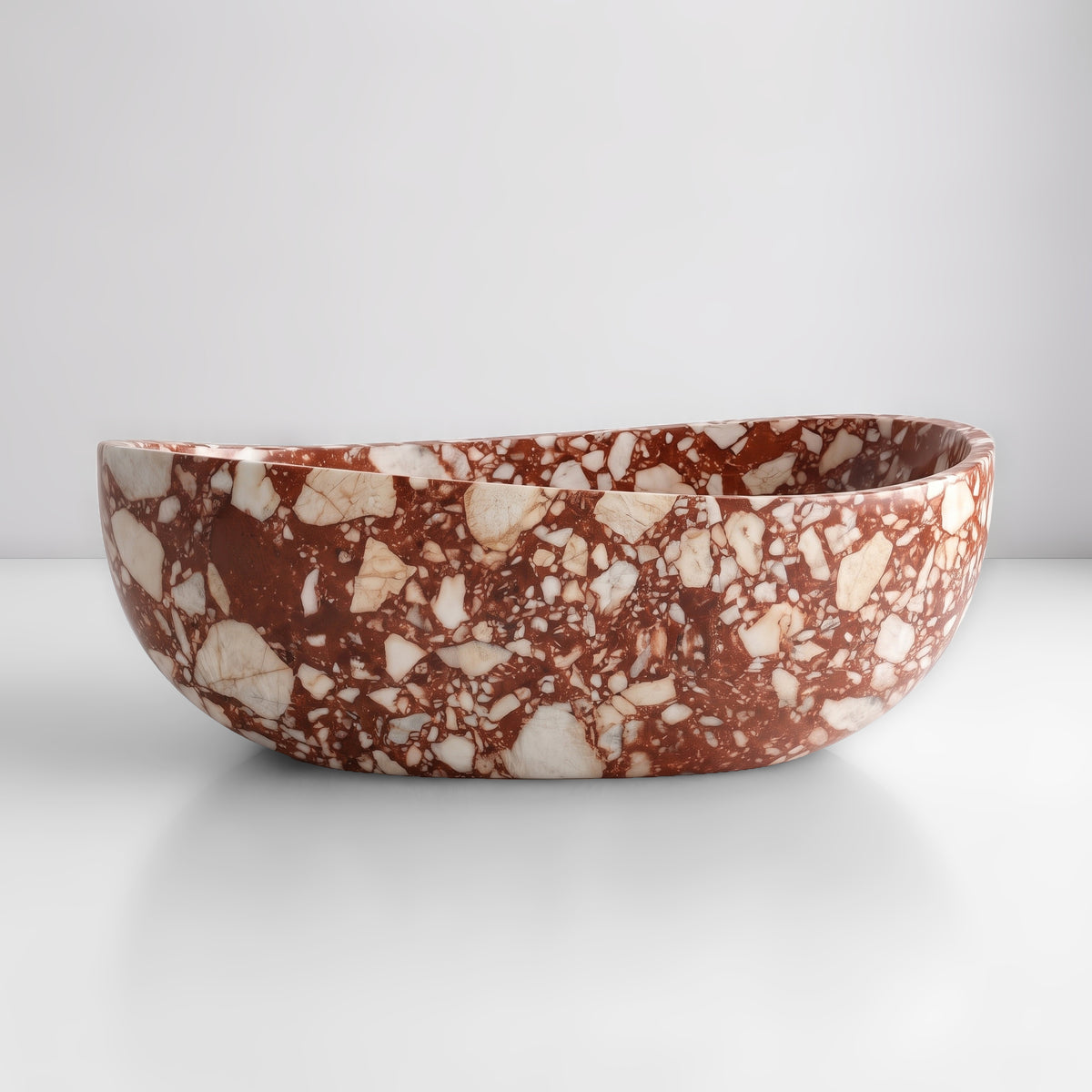 Front studio view of the freestanding Red Bulgari marble wave bathtub, showcasing the organic dipping rim silhouette and bold terracotta-red breccia ground embedded with cream and white calcite clasts across the exterior.