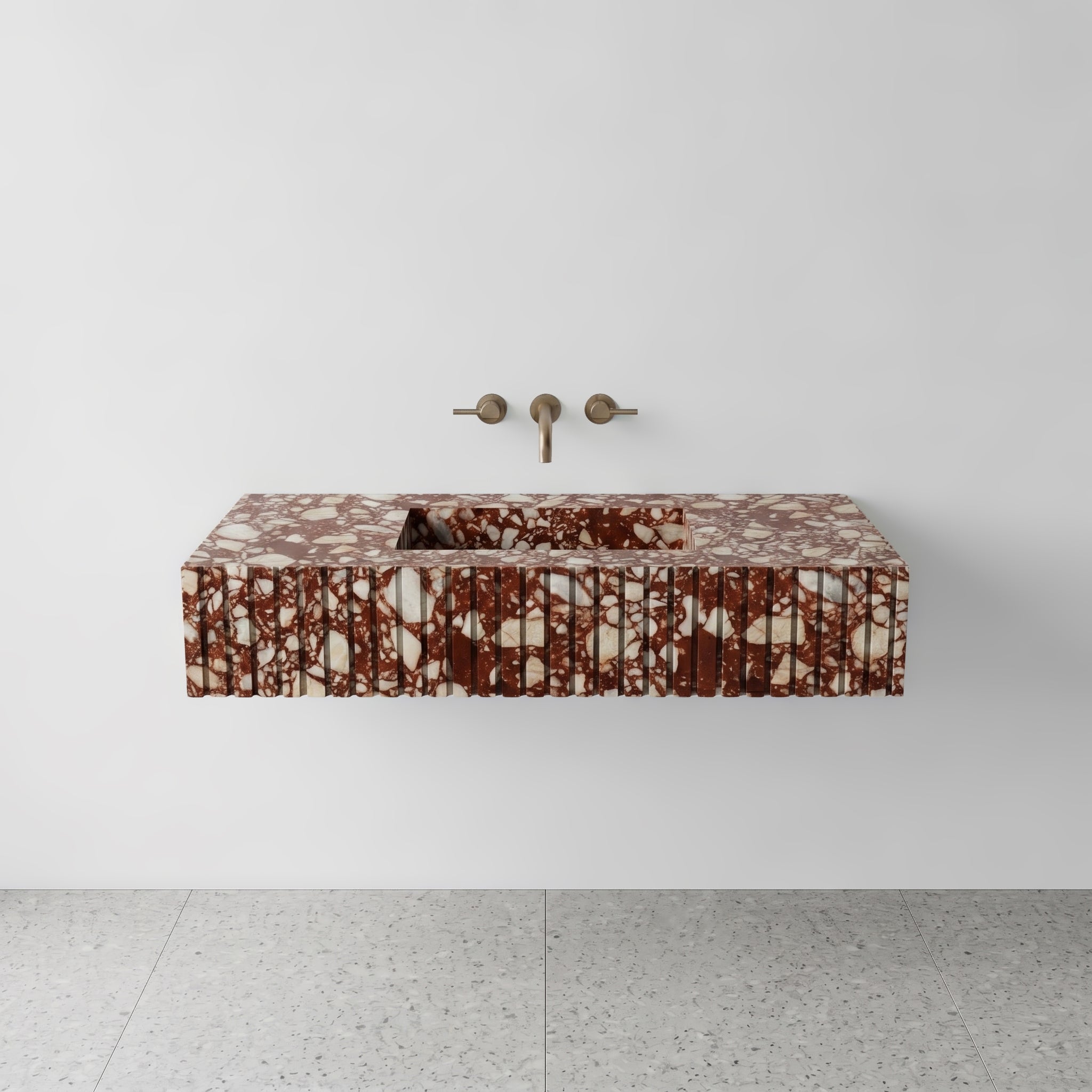 Front studio view of a luxury floating single washbasin carved from brecciated Red Bulgari marble, featuring an architectural fluted apron front design and a wall-mounted brushed bronze faucet.