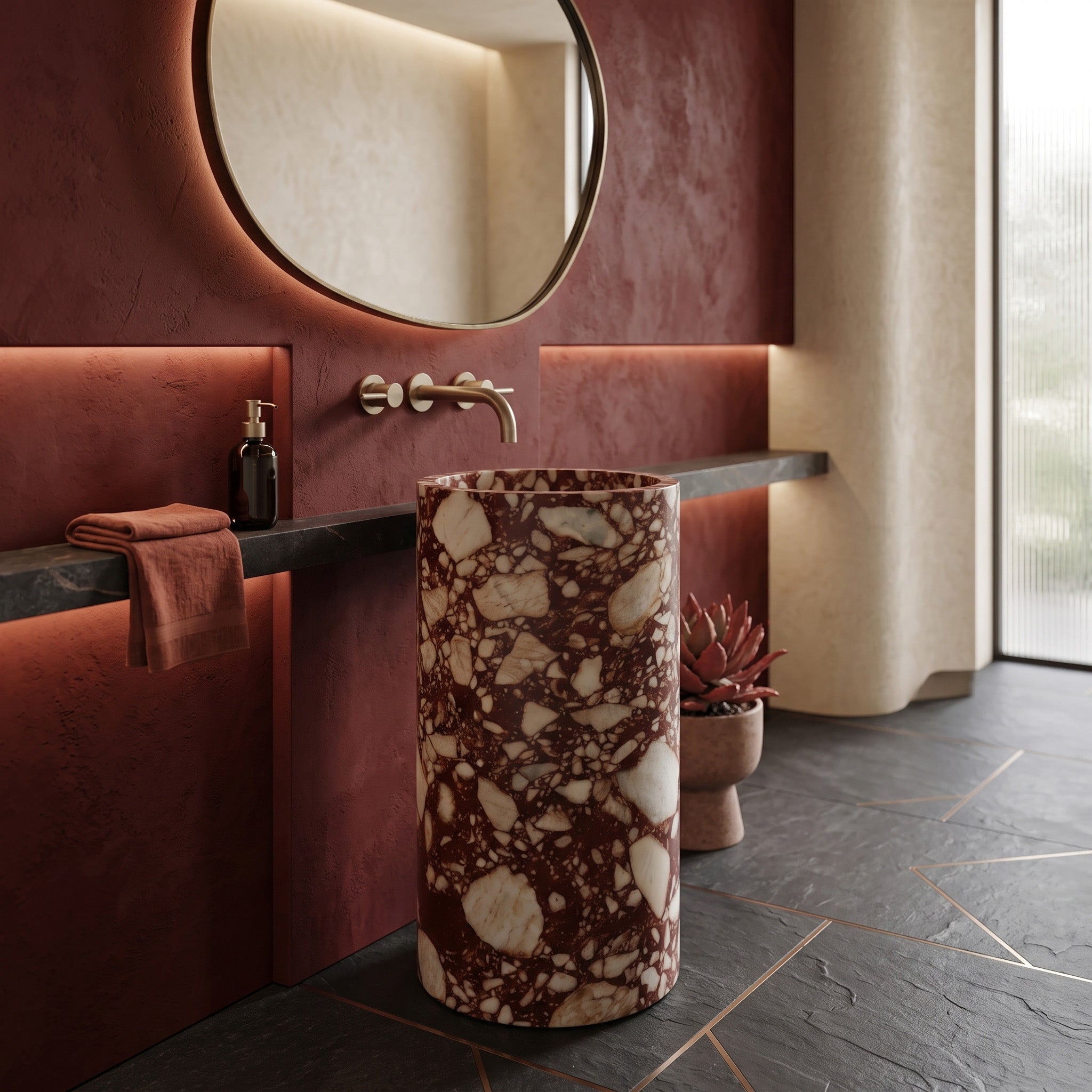 Freestanding Red Bulgari marble round pedestal basin in a luxury powder room with deep red plaster walls, backlit organic mirror, and black slate flooring.