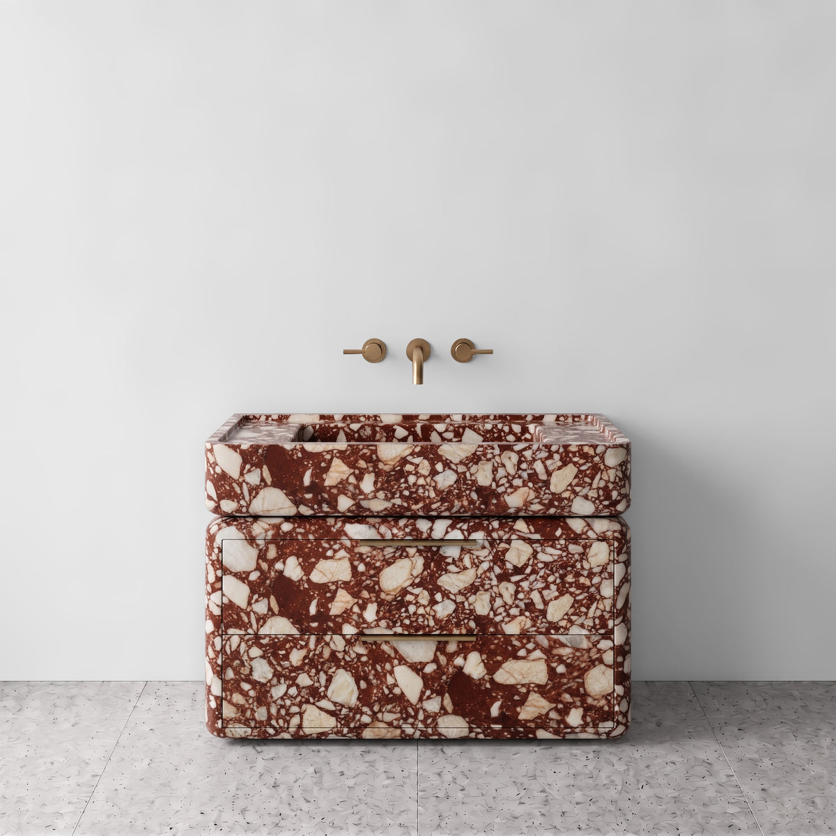 Front studio view of the freestanding Red Bulgari marble double-drawer vanity, featuring rounded corners, an integrated trough basin, horizontal brushed brass pulls, and wall-mounted three-hole brass tapware set against a white wall and terrazzo flooring.