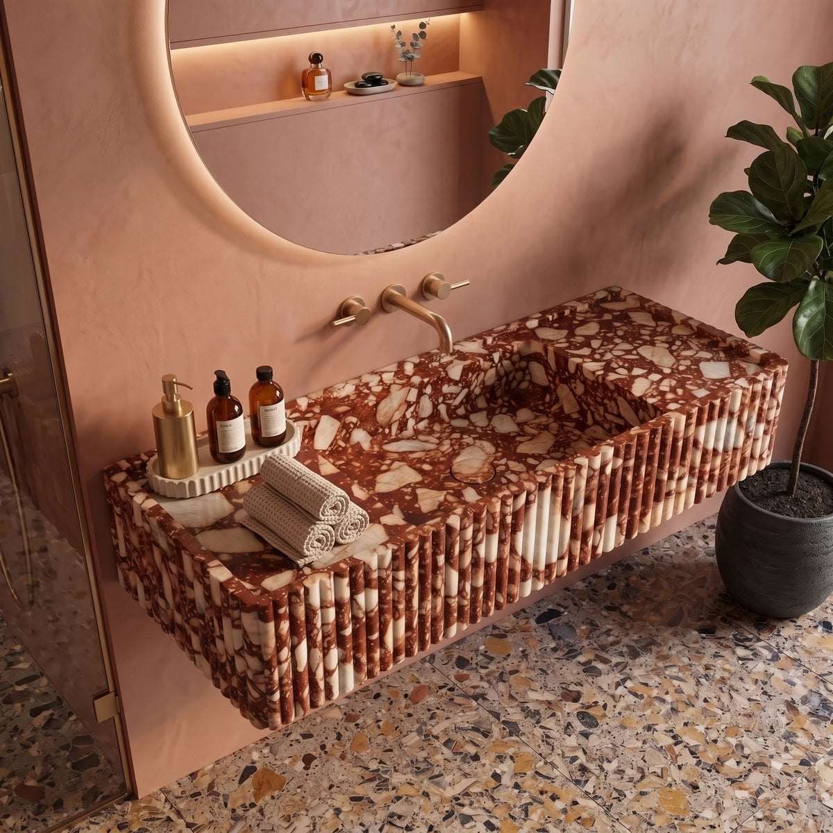 Warm contemporary bathroom interior featuring the floating Red Bulgari fluted stone sink paired with terracotta plaster walls, a backlit round mirror, a potted plant, and terrazzo flooring.