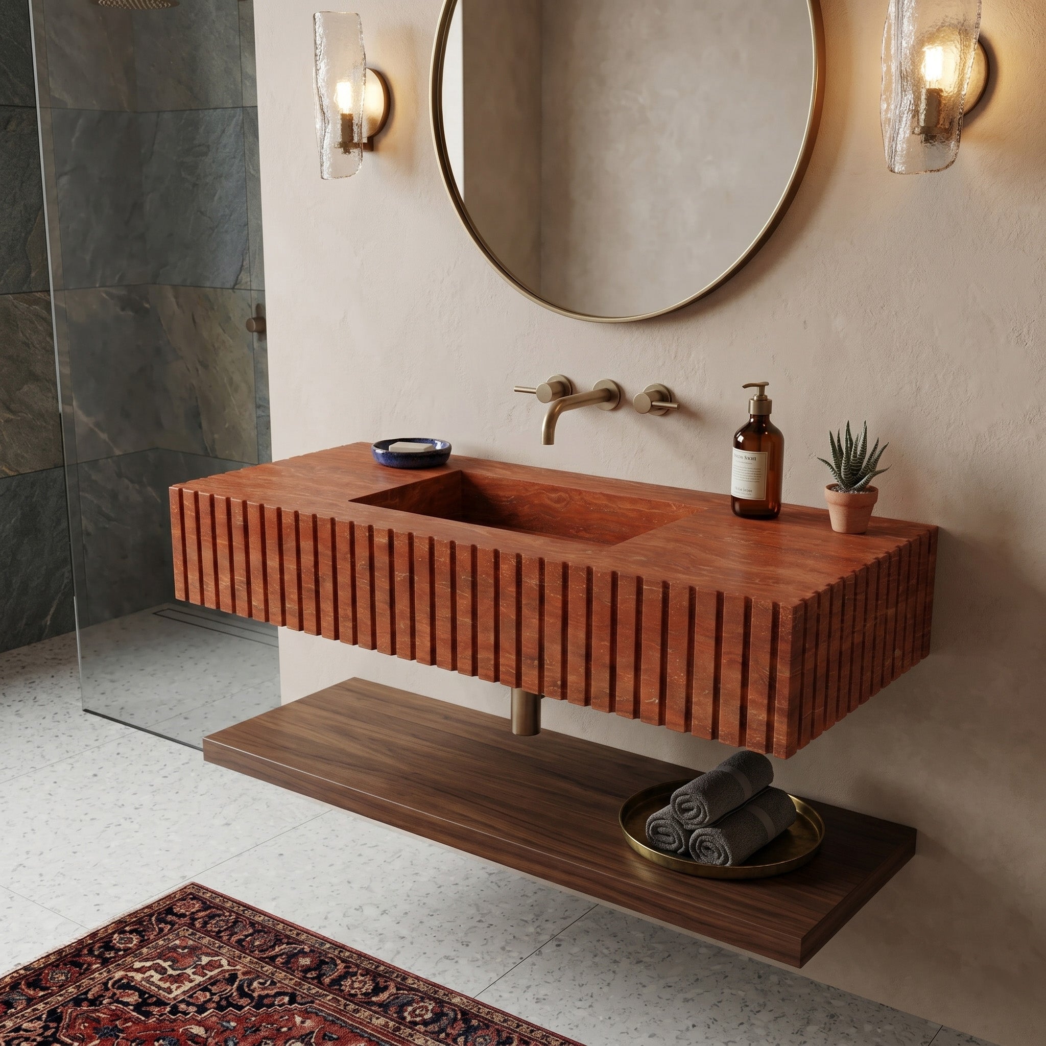 Mediterranean organic bathroom interior featuring a fluted red travertine single sink, a floating dark wood shelf, a round brass mirror, textured plaster walls, and an antique red patterned rug.