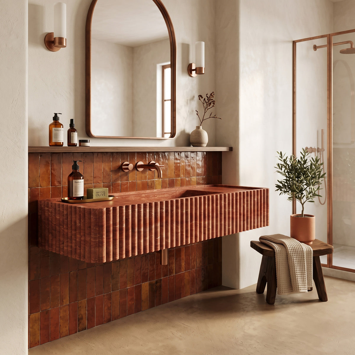 Warm Mediterranean bathroom interior featuring the floating Red Travertine fluted stone sink set against glossy caramel zellige tiles, an arched timber mirror, and copper shower framing.