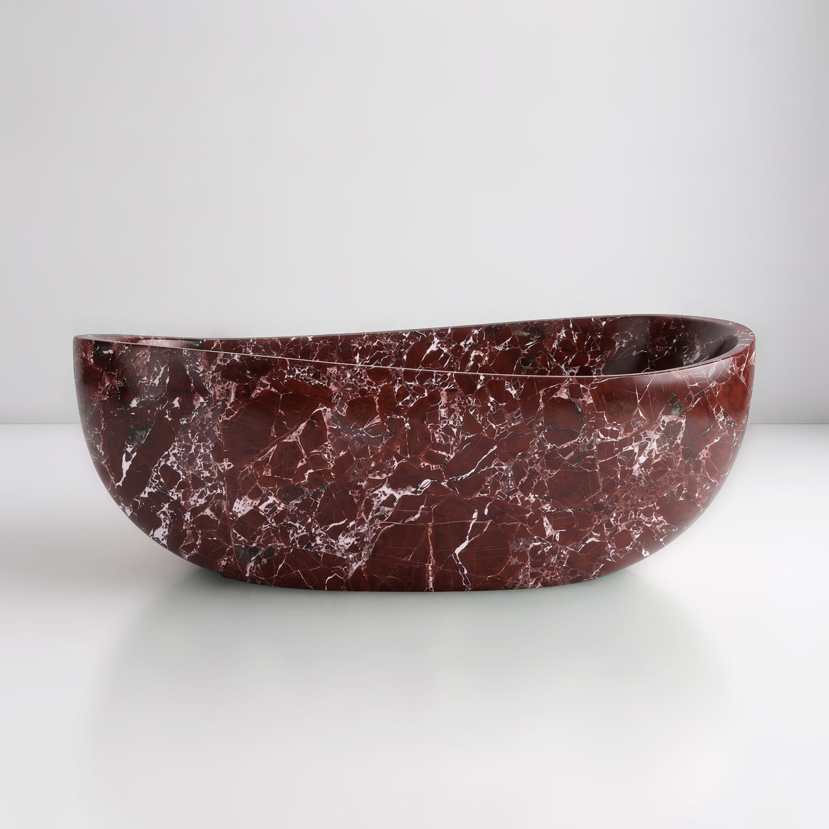 Front studio view of the freestanding Rosso Levanto marble wave bathtub, showcasing the organic dipping rim silhouette and deep cherry-red stone patterned with vivid white calcite spiderweb veining across the exterior.