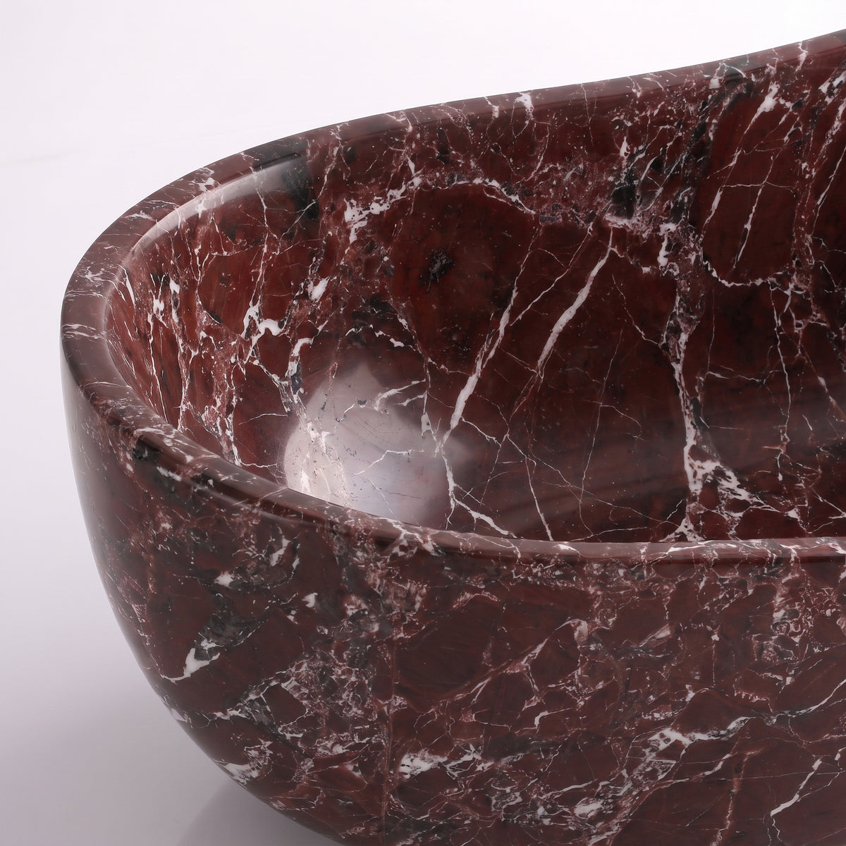 Close-up overhead detail shot of the Rosso Levanto marble wave bathtub rim, displaying the hand-finished curve, polished stone texture, and crisp white calcite veins traversing deep plum-red marble.