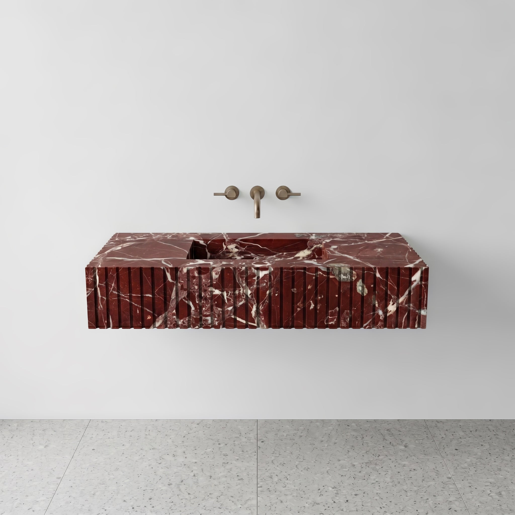 Front studio view of a luxury floating single sink vanity carved from deep red Rosso Levanto marble, showcasing a geometric fluted apron front design and a wall-mounted brushed bronze faucet.