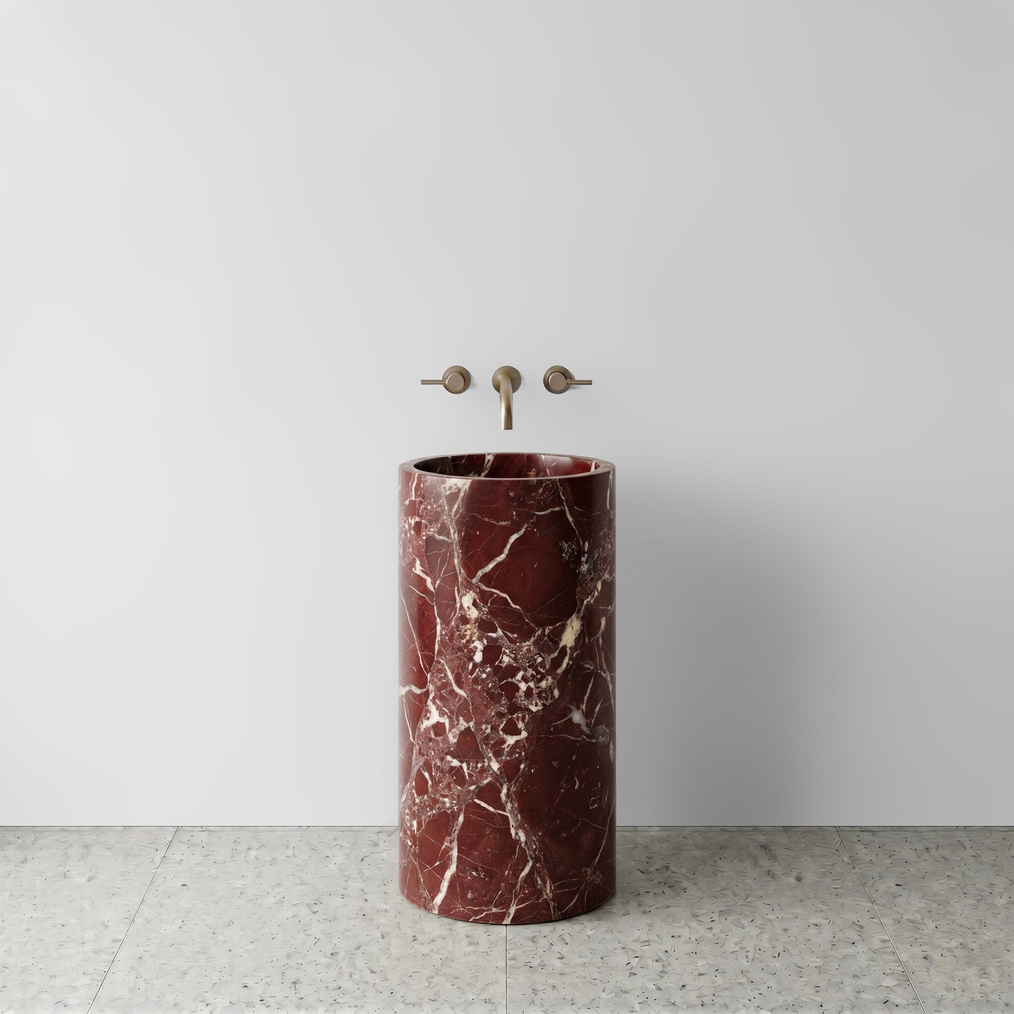 Front view of a monolithic Rosso Levanto red marble cylinder pedestal sink on light terrazzo flooring with wall-mounted bronze tapware.
