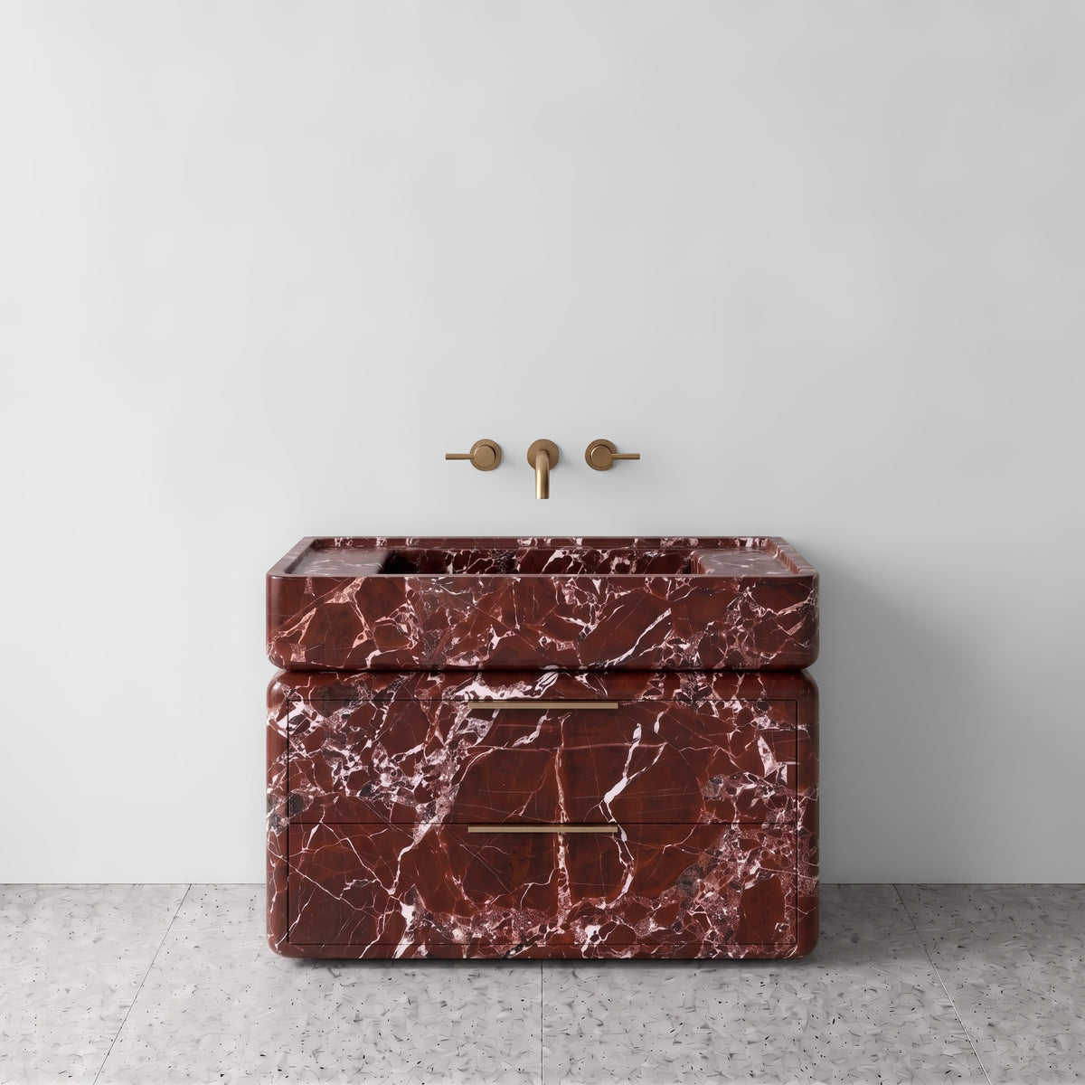Front studio view of the freestanding Rosso Levanto marble double-drawer vanity, featuring rounded corners, an integrated trough basin, horizontal brushed brass pulls, and wall-mounted three-hole brass tapware set against a white wall and terrazzo flooring.