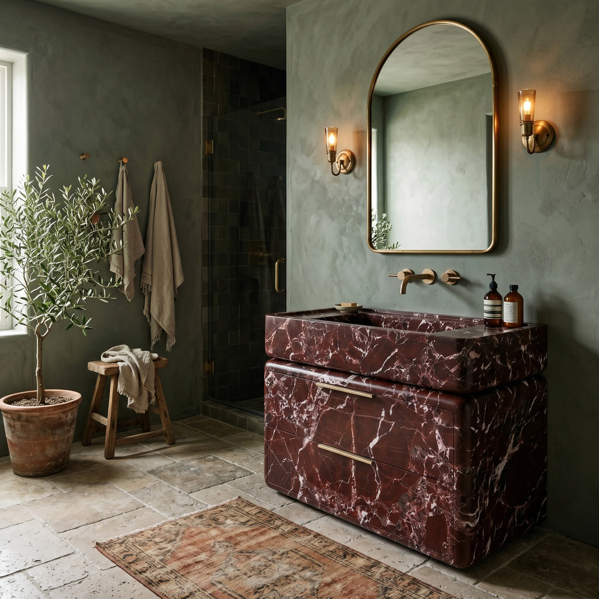 Atmospheric luxury bathroom interior featuring the curved Rosso Levanto marble double-drawer vanity paired with wall-mounted brass tapware, an arched brass mirror flanked by glowing sconces, sage-green limewash walls, a potted olive tree, and a weathered vintage runner on stone tile flooring.