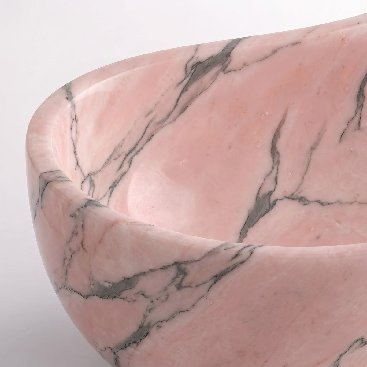 Close-up overhead detail shot of the Rosso Portogallo marble wave bathtub rim, displaying the hand-finished curve, polished stone surface, and fine charcoal veining traversing soft pink marble.