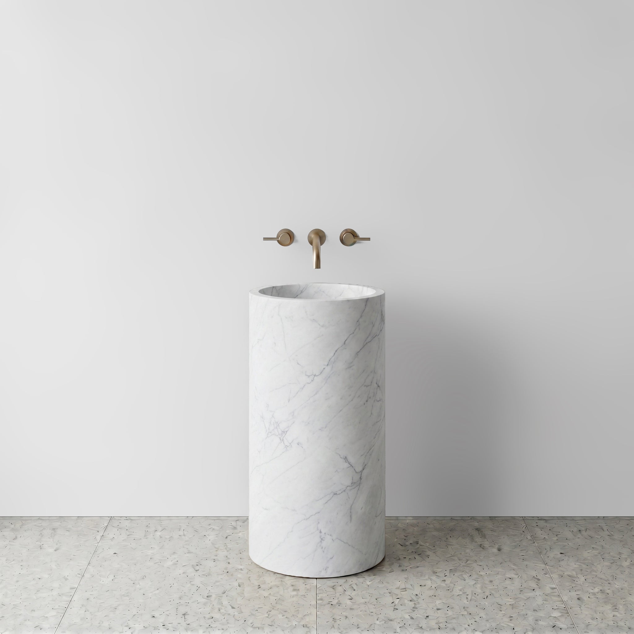 Front view of a solid White Carrara marble cylinder pedestal sink on light terrazzo flooring with wall-mounted bronze tapware.