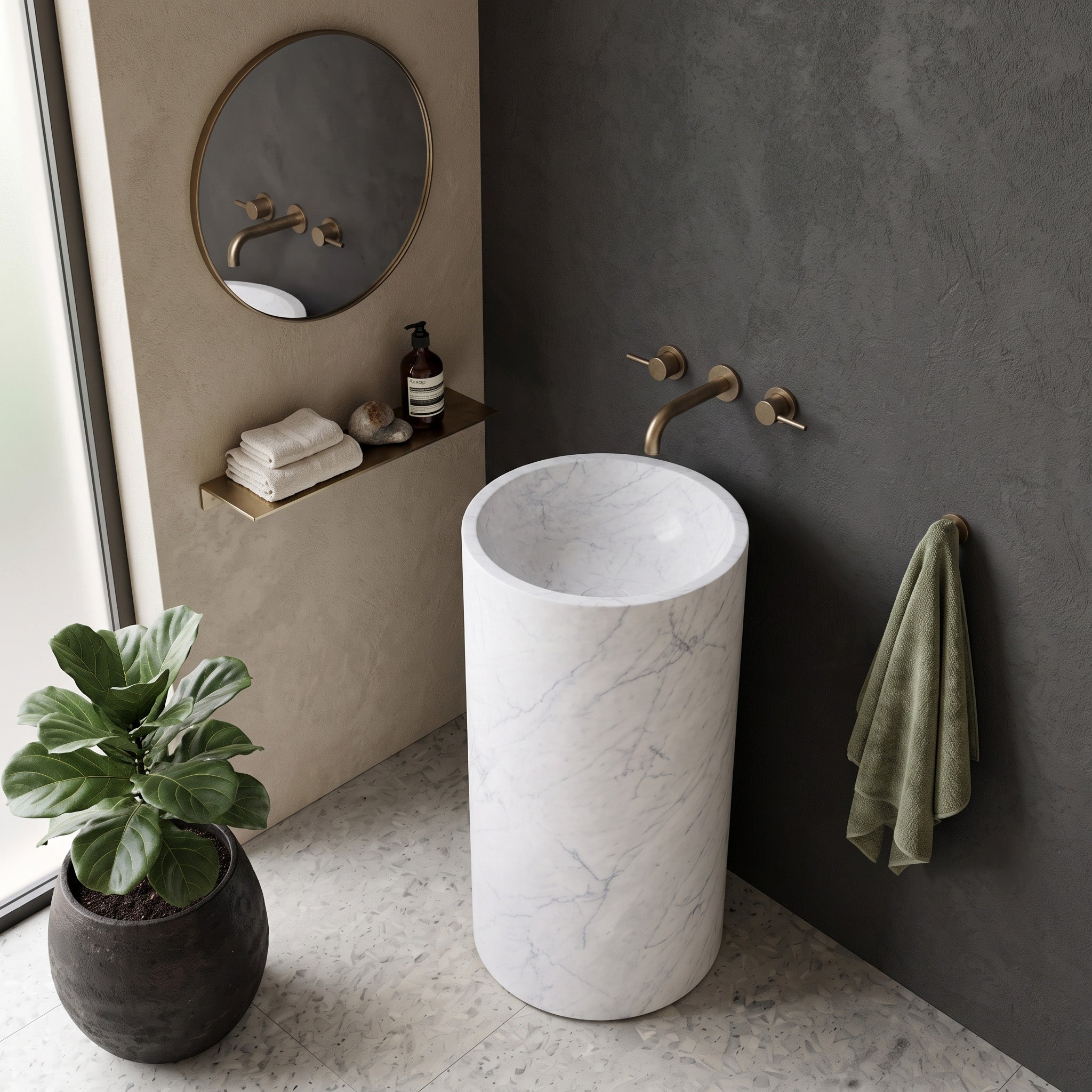 Freestanding White Carrara marble round pedestal basin in a minimalist bathroom with grey concrete walls, marble shelf, and organic brass mirror.