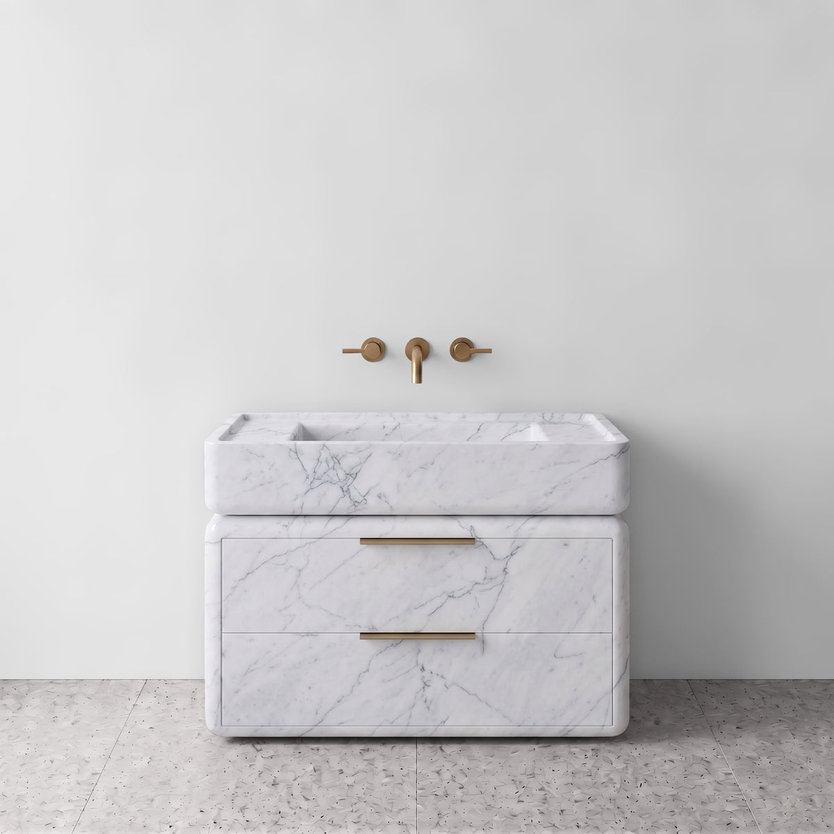Front studio view of the freestanding White Carrara marble double-drawer vanity, featuring rounded corners, an integrated trough basin, horizontal brushed brass pulls, and wall-mounted three-hole brass tapware set against a white wall and terrazzo flooring.